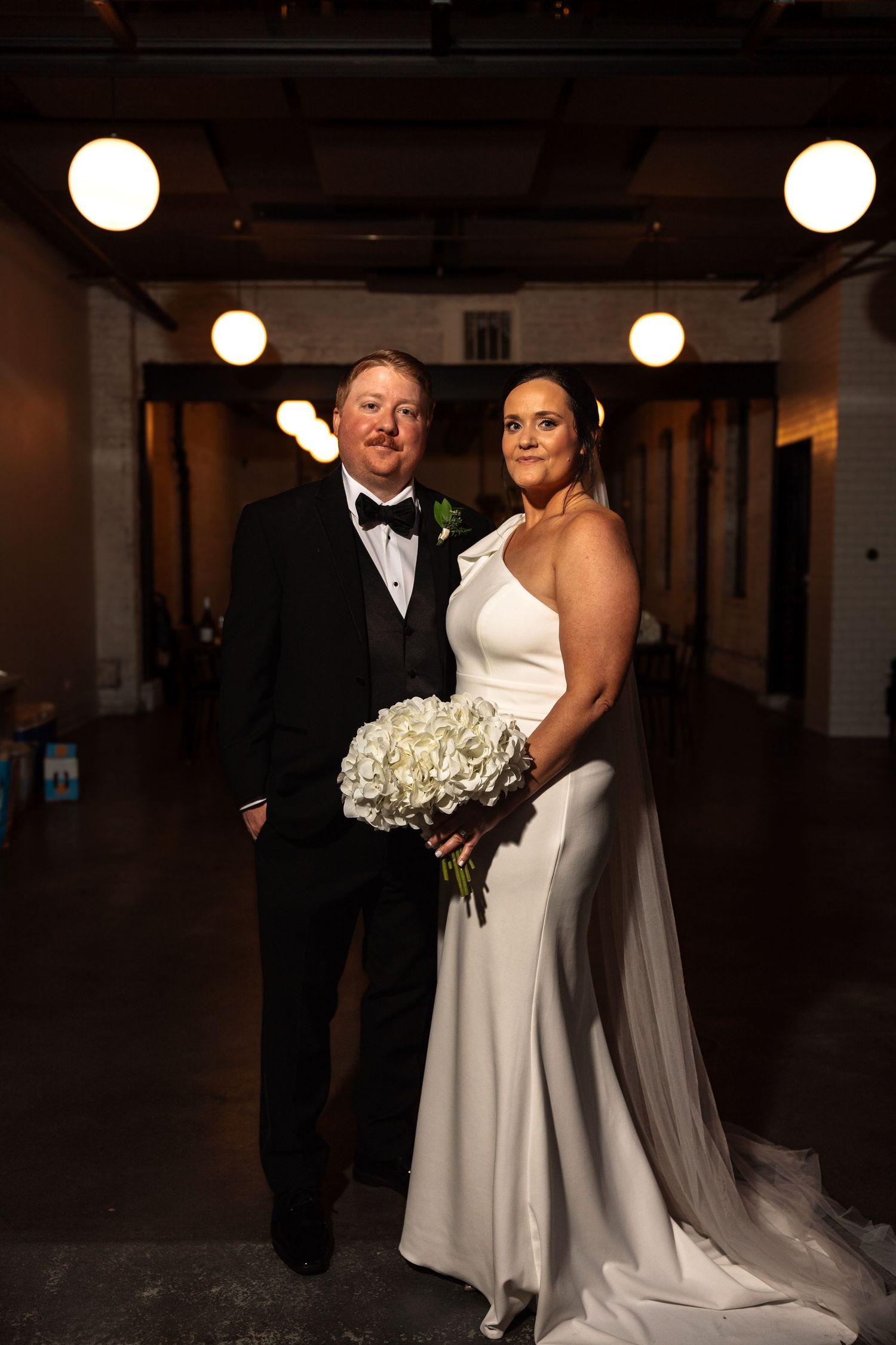 Bride and groom posing in industrial venue, bride in white gown holding white hydrangea bouquet.