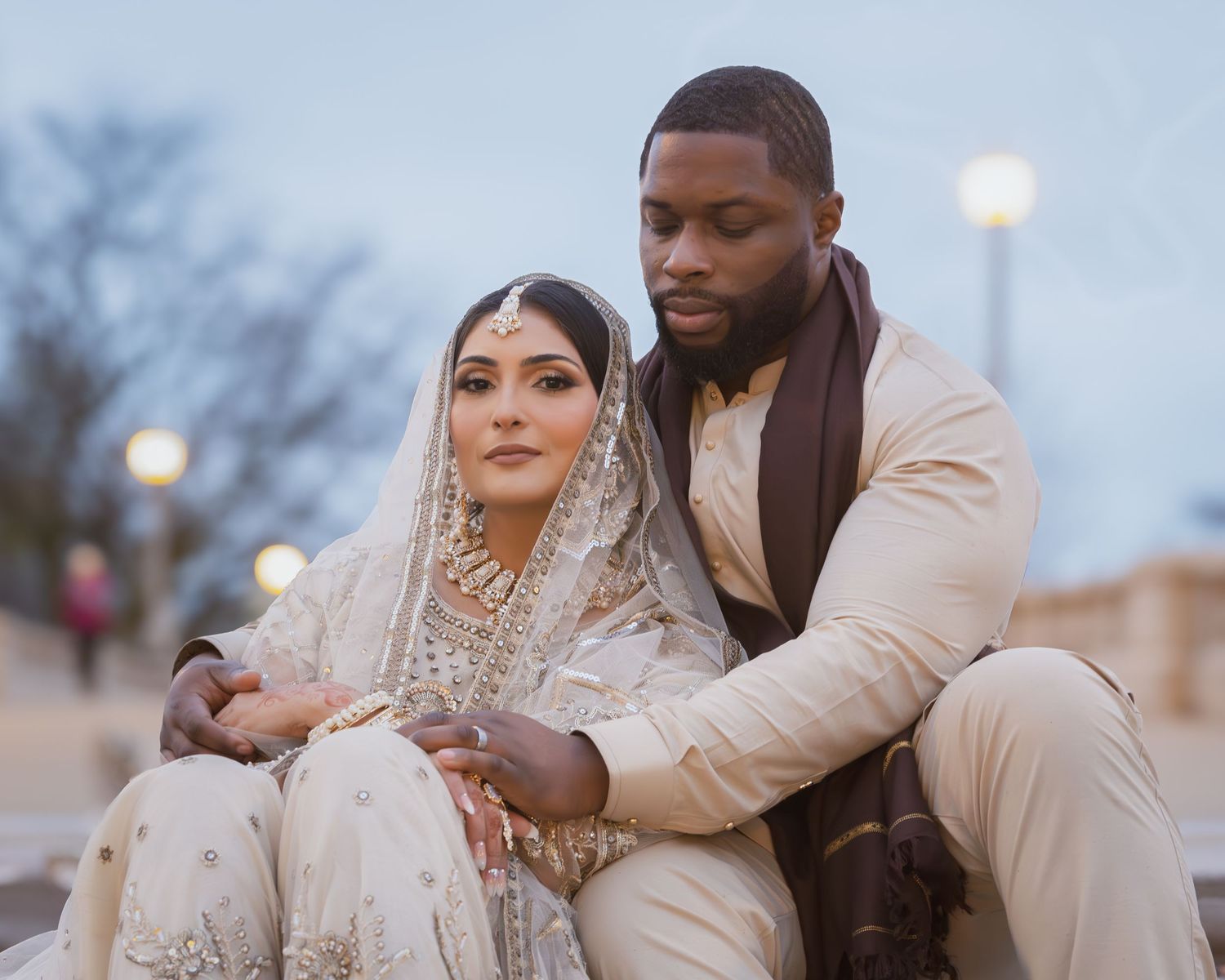 South Asian bride in white embroidered bridal lehenga sits closely with groom in cream sherwani.