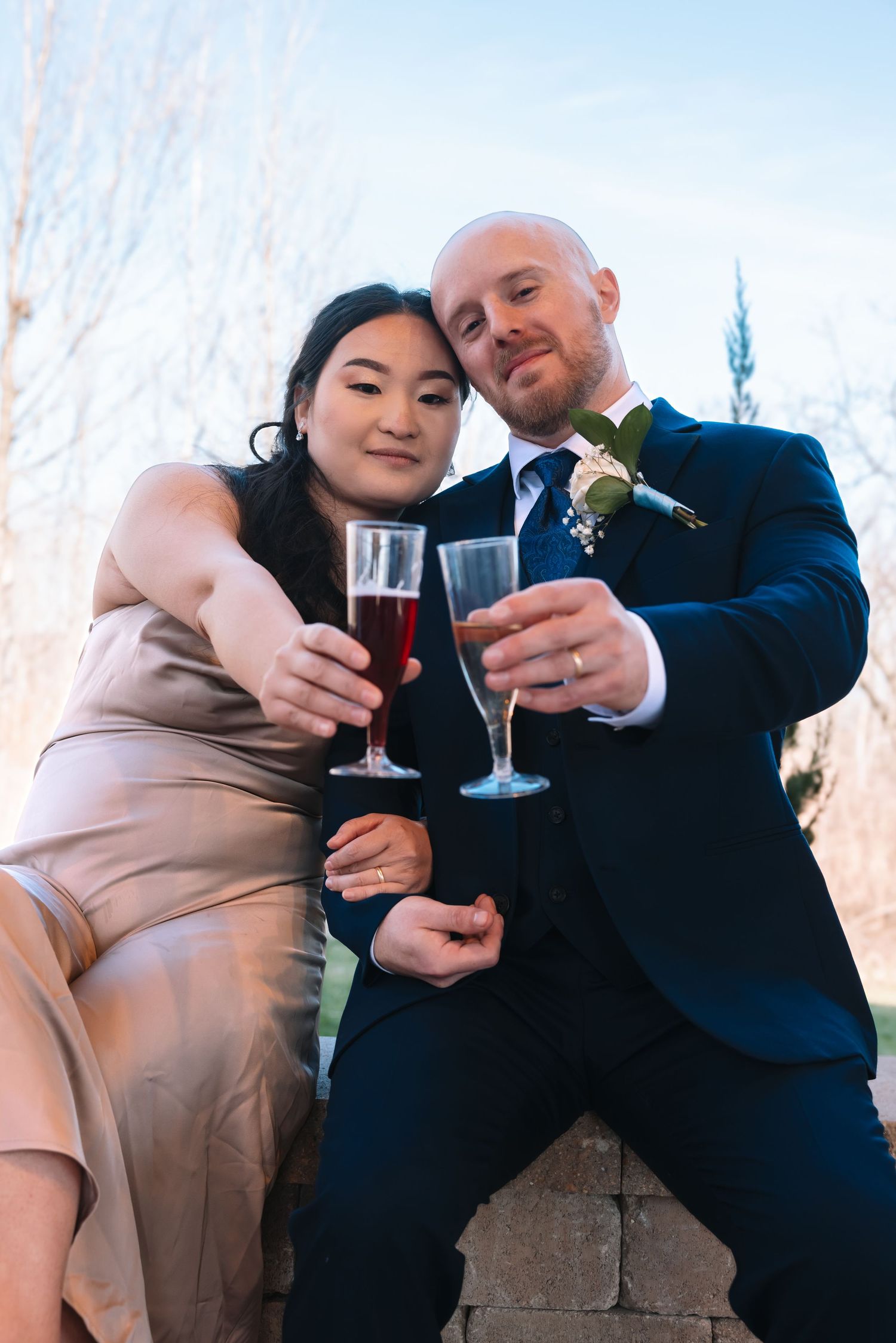 Bride and groom in navy suit toasting with red champagne glasses at their outdoor wedding celebration.