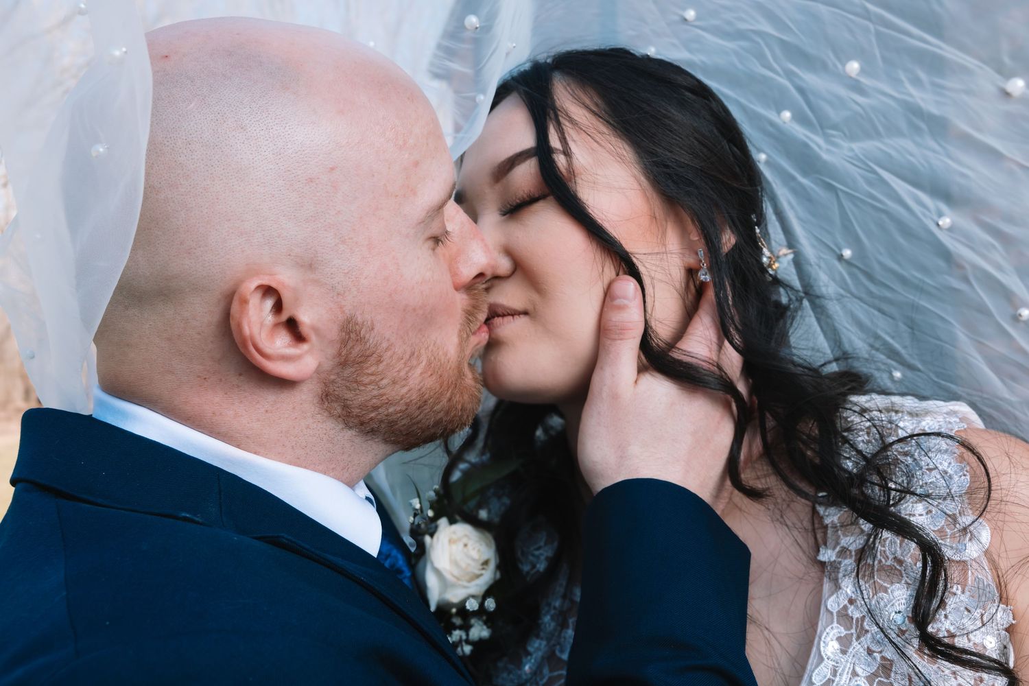 Bride and groom sharing a romantic kiss under a sheer wedding veil, groom in navy suit cradling brides face.