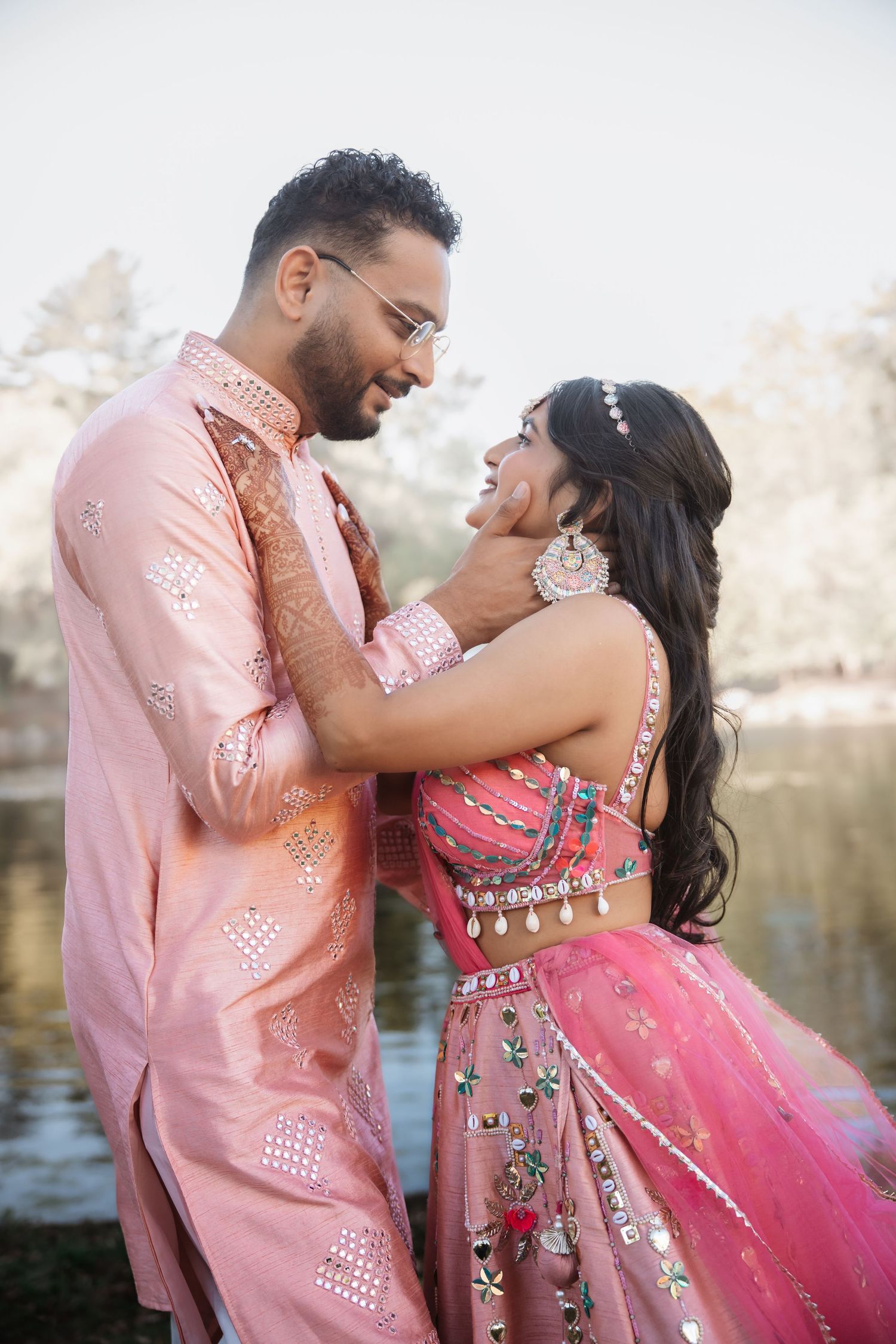 Indian couple in pink traditional wedding outfits sharing a tender moment by lake.