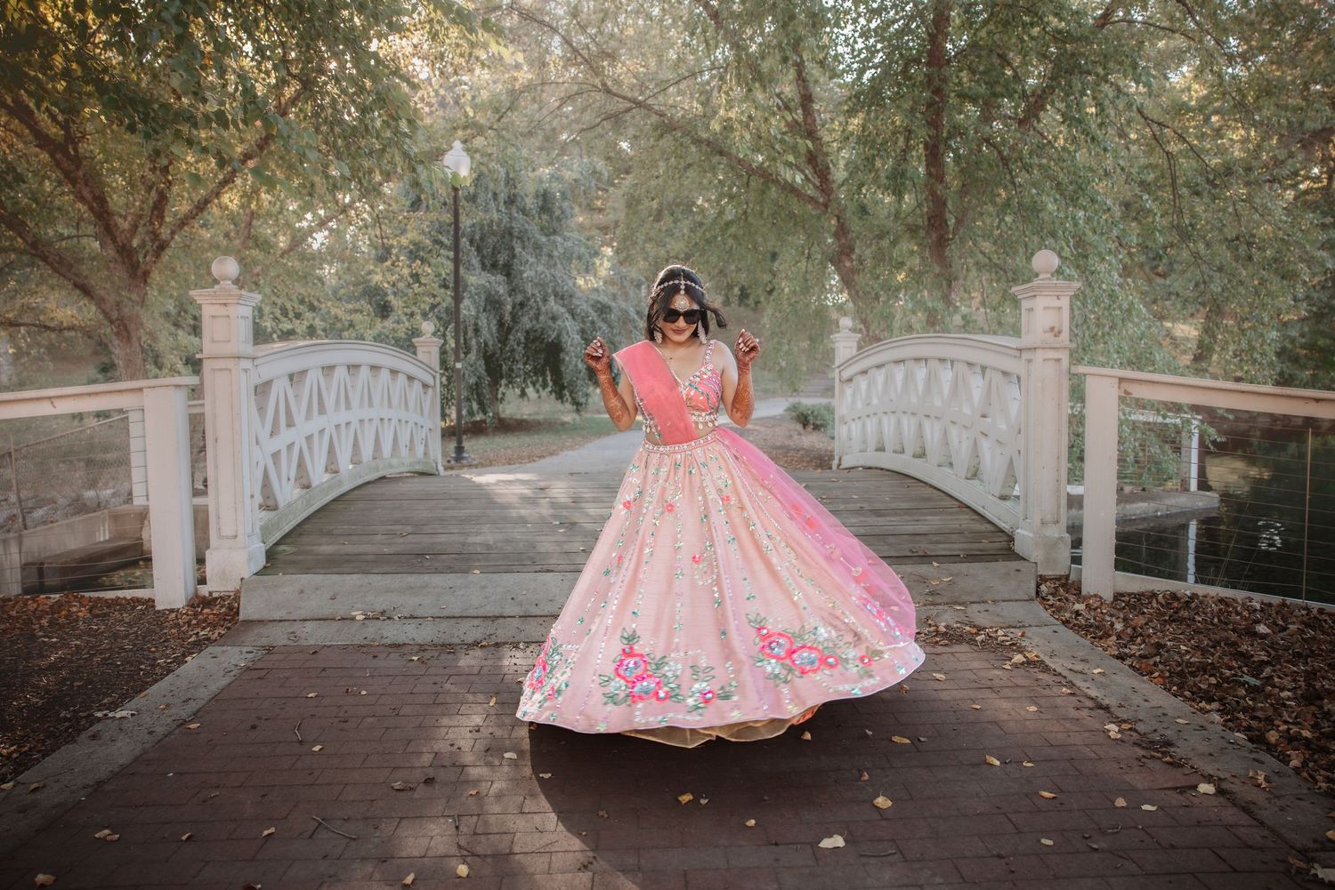 Indian bride twirling in pink embroidered lehenga on wooden bridge surrounded by trees.