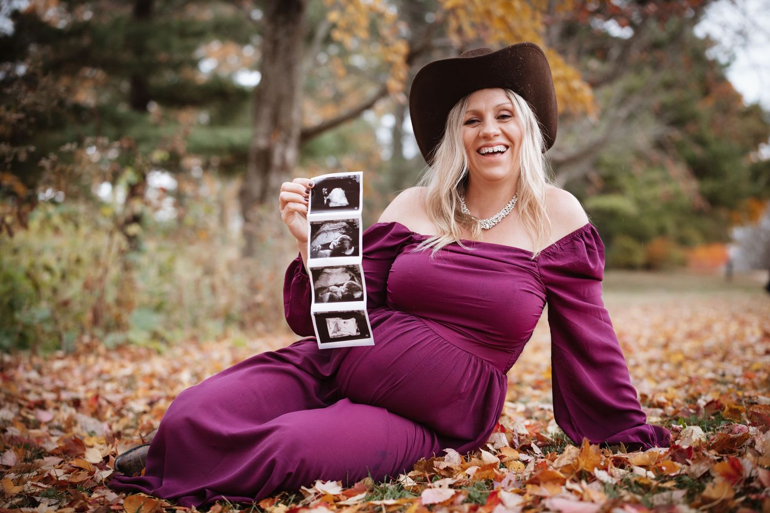 Laughing pregnant woman in purple dress and cowboy hat sitting in fall leaves holding ultrasound images.