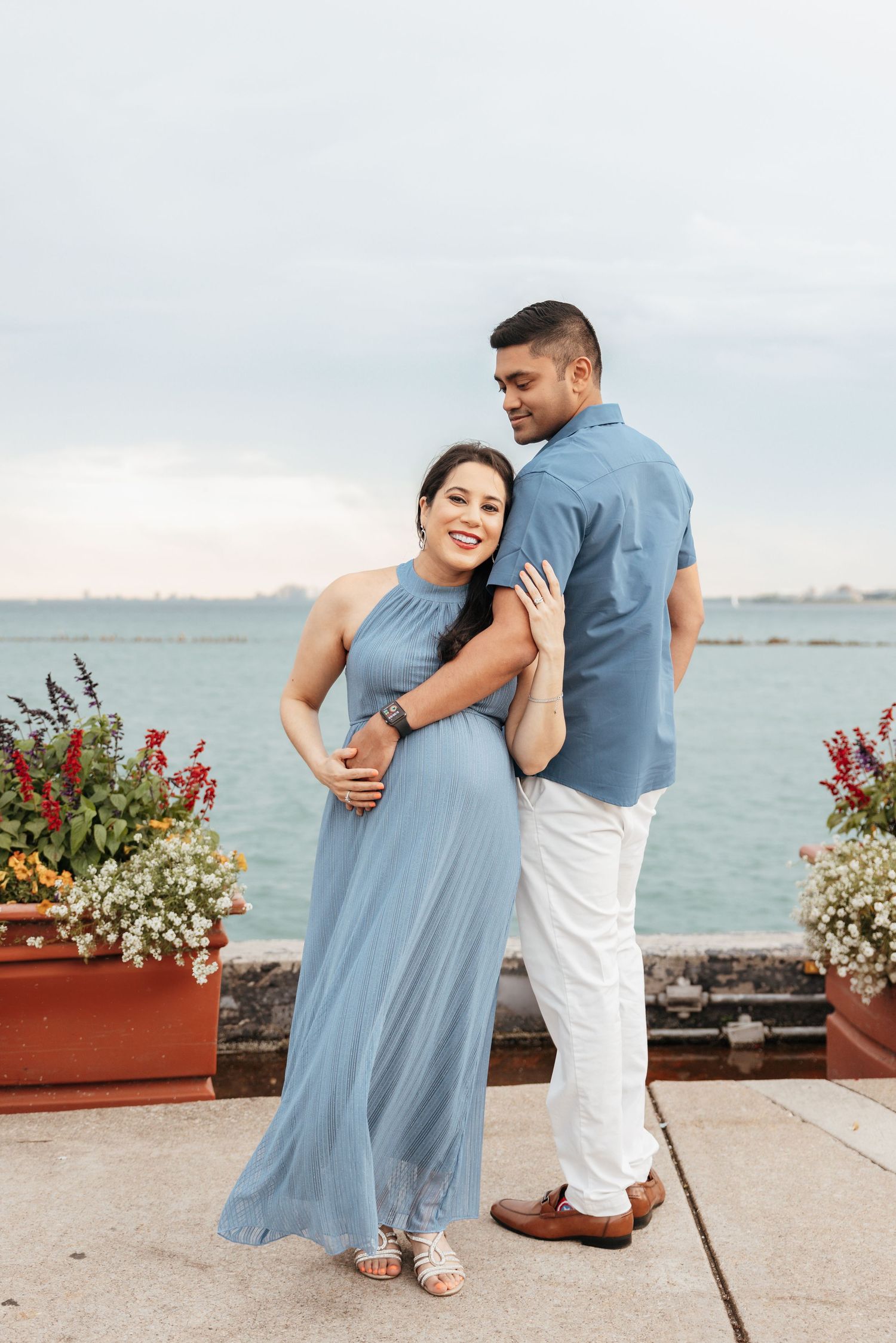 Pregnant couple in coordinating blue outfits posing by colorful flowers near water.