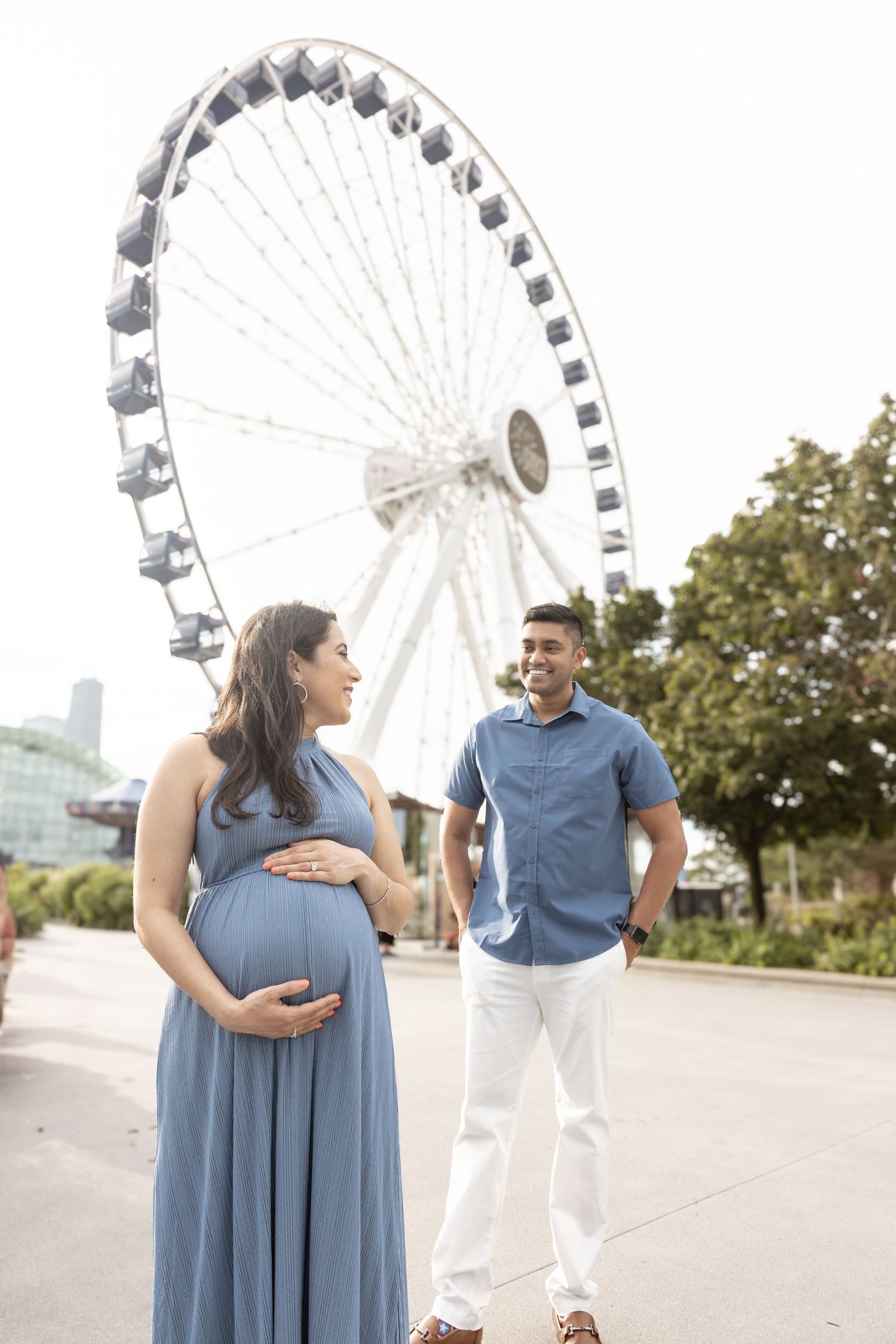 Pregnant woman in blue dress with partner near Ferris wheel.