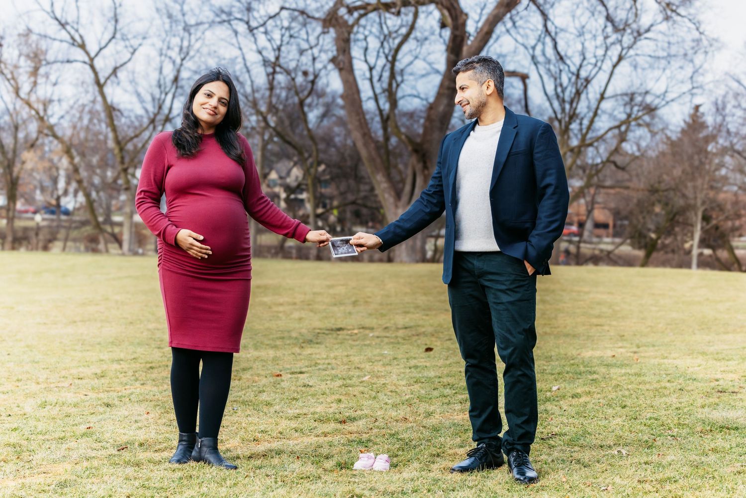 Pregnant woman in red dress and man in navy blazer hold ultrasound photo in a park.