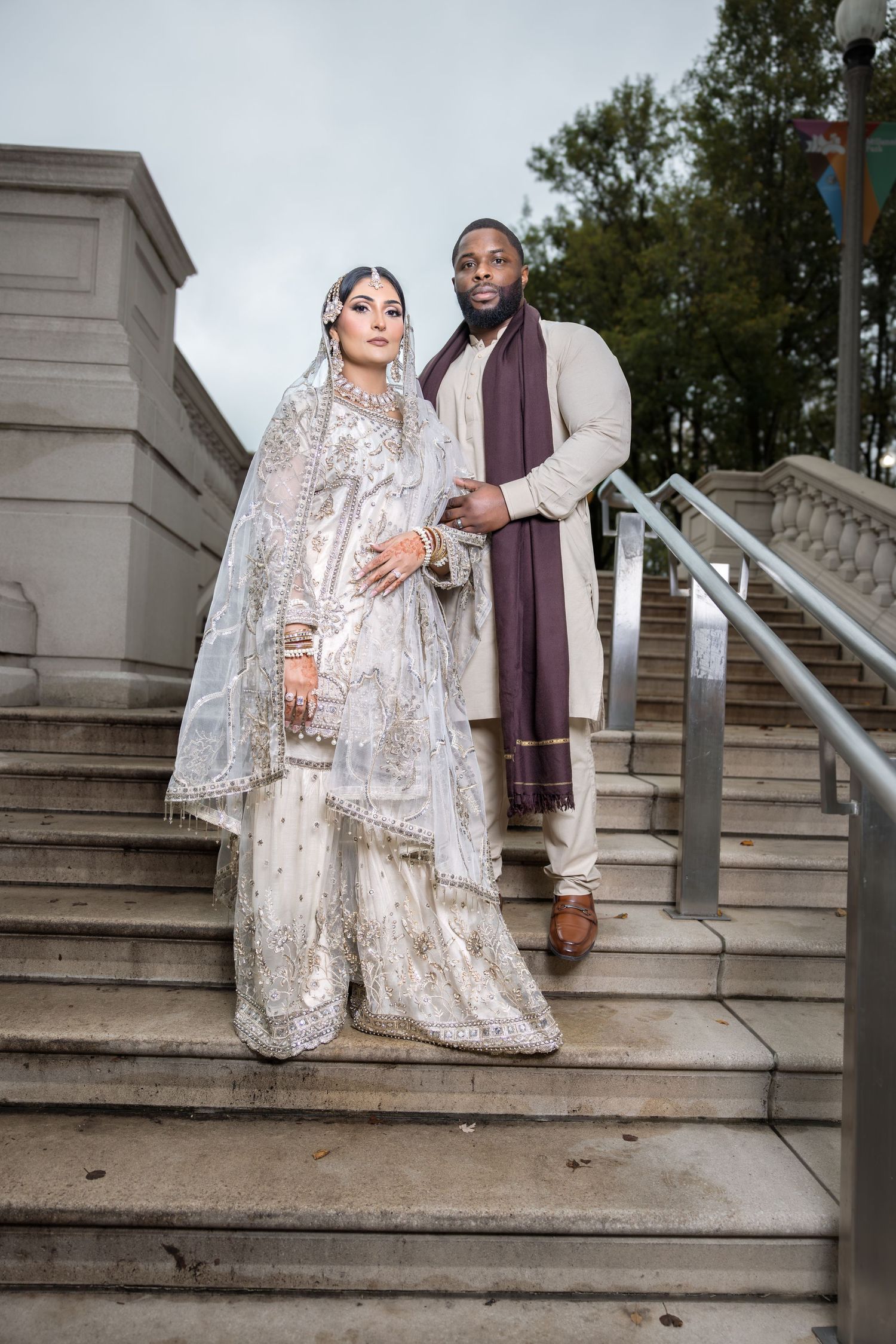 Couple in traditional South Asian Muslim wedding outfits posing on stone steps with trees behind.