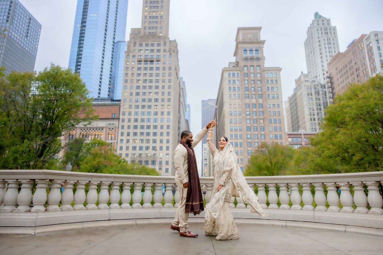 Groom twirls bride in white embroidered dupatta on elegant bridge with Chicago skyscrapers behind.