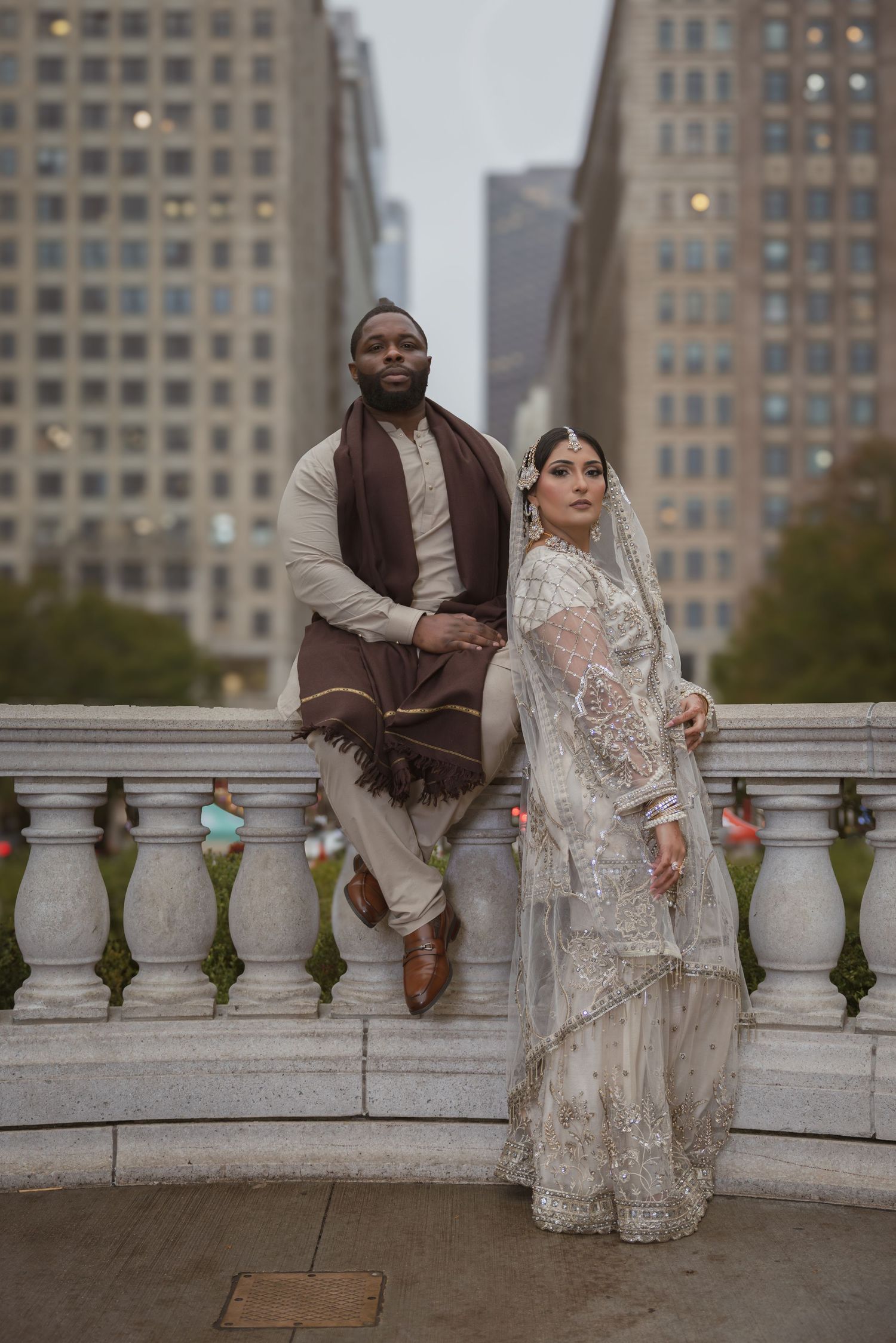 South Asian bride and groom in traditional wedding attire pose elegantly on a stone balcony in the city.