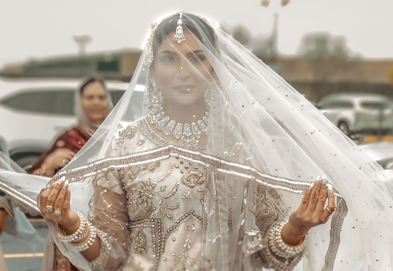 A South Asian bride in white embroidered lehenga holds her veil open outdoors on her wedding day.