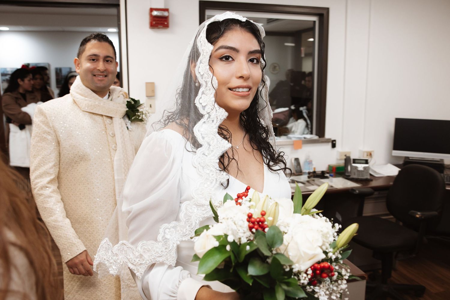 Radiant bride in white lace veil holds bouquet of white roses while groom in ivory sherwani stands behind her.