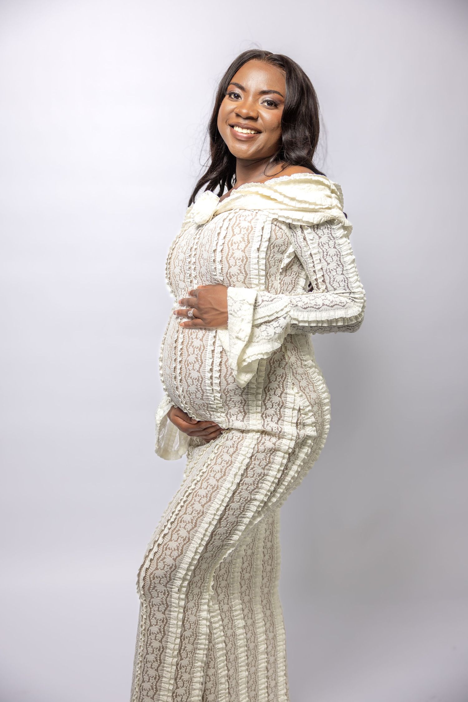 Glowing pregnant Black woman in cream textured lace dress smiling while cradling her baby bump.