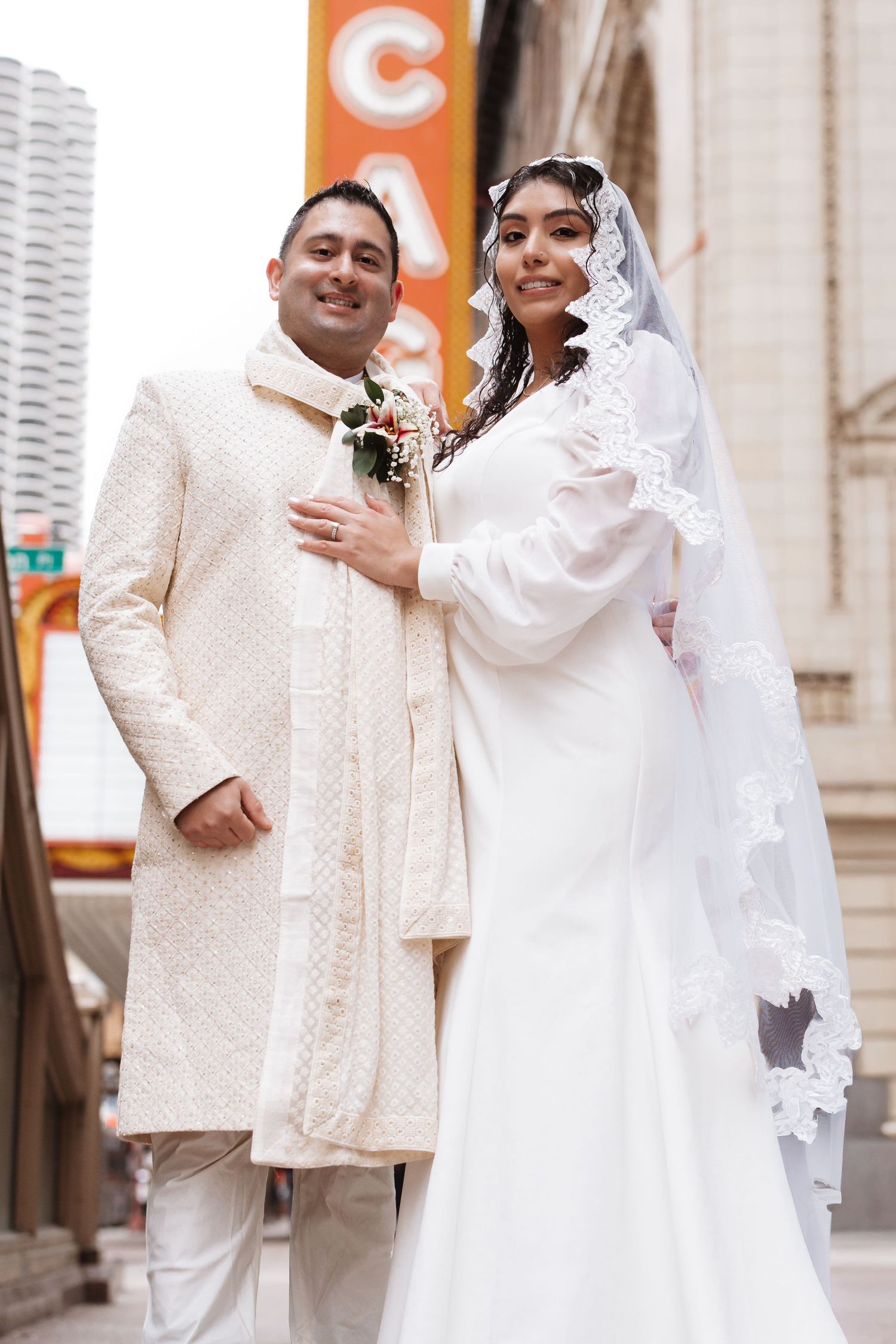 Bride and groom in elegant white wedding attire pose on Chicago city street near theater signage.