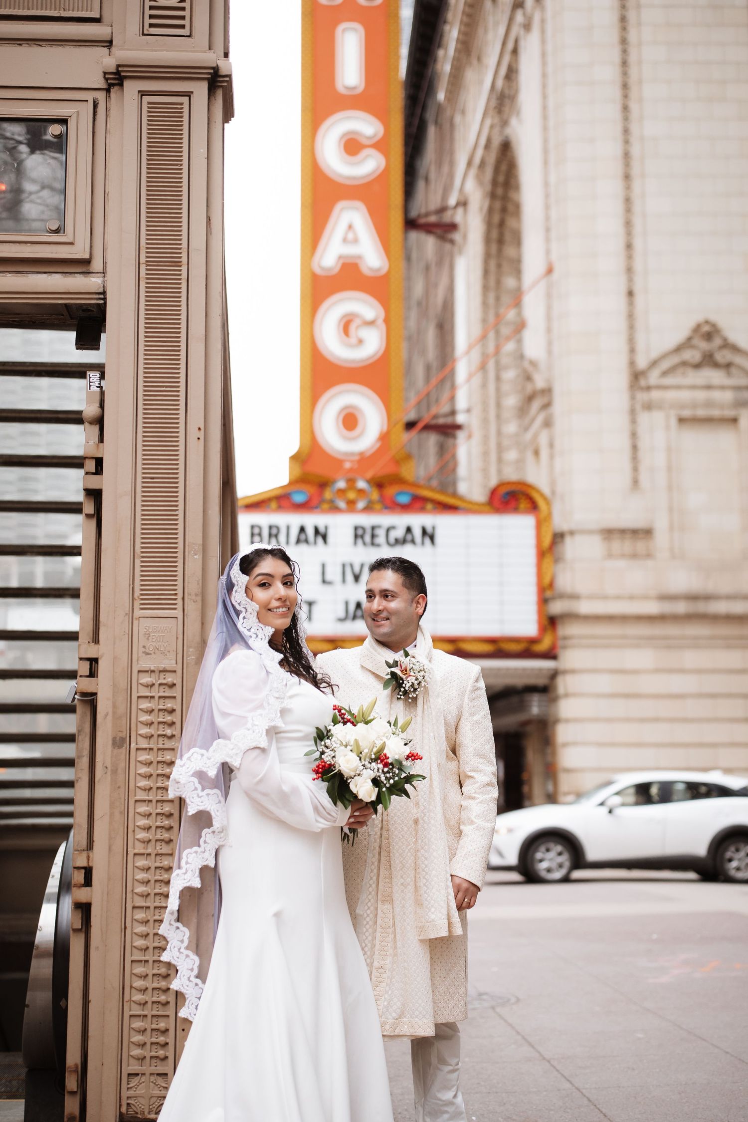Bride and groom stand together outside Chicago Theatre, bride holding bouquet of flowers.