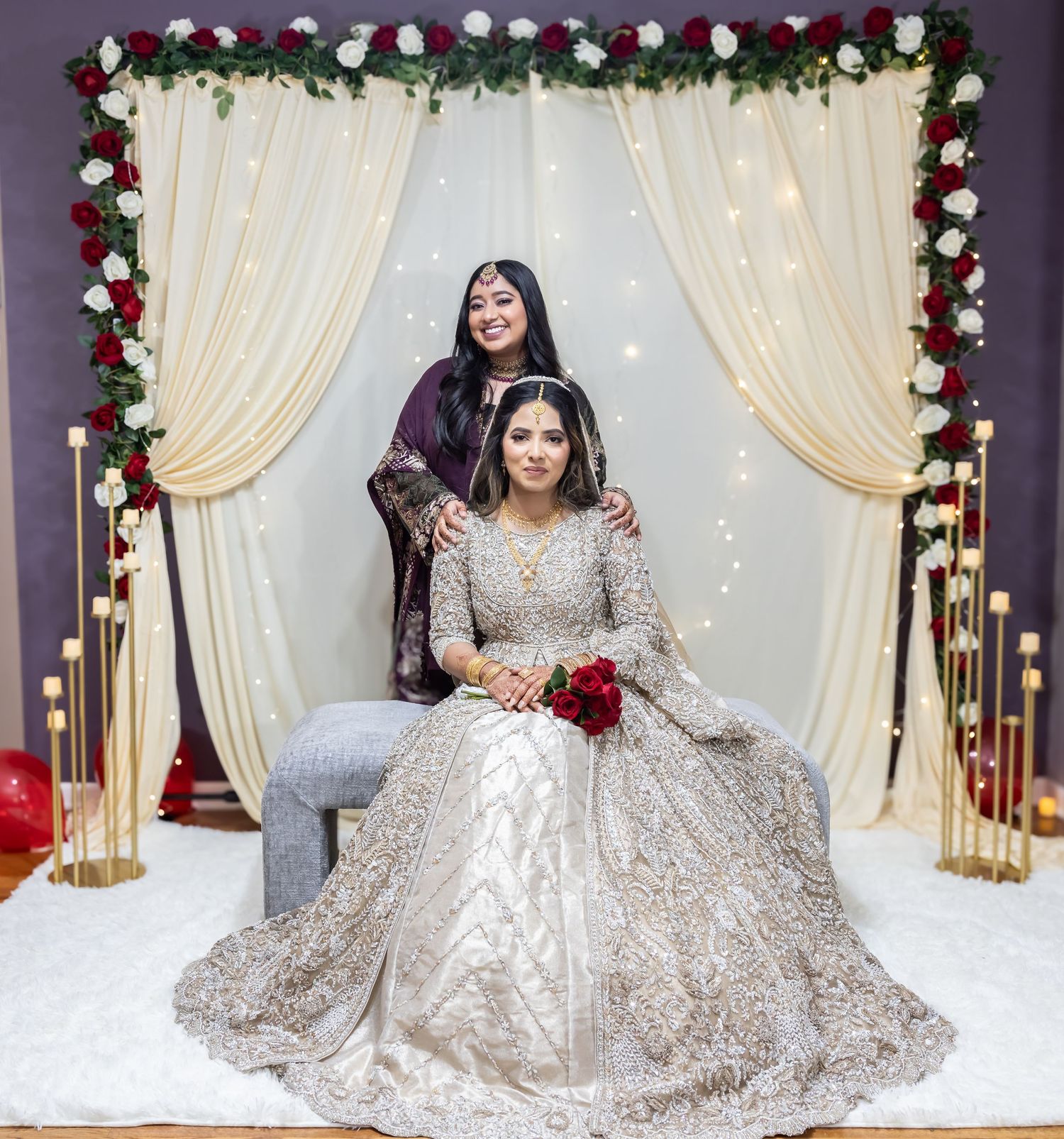 Bride in gold lehenga sits holding red roses while woman in purple stands behind at floral wedding arch.