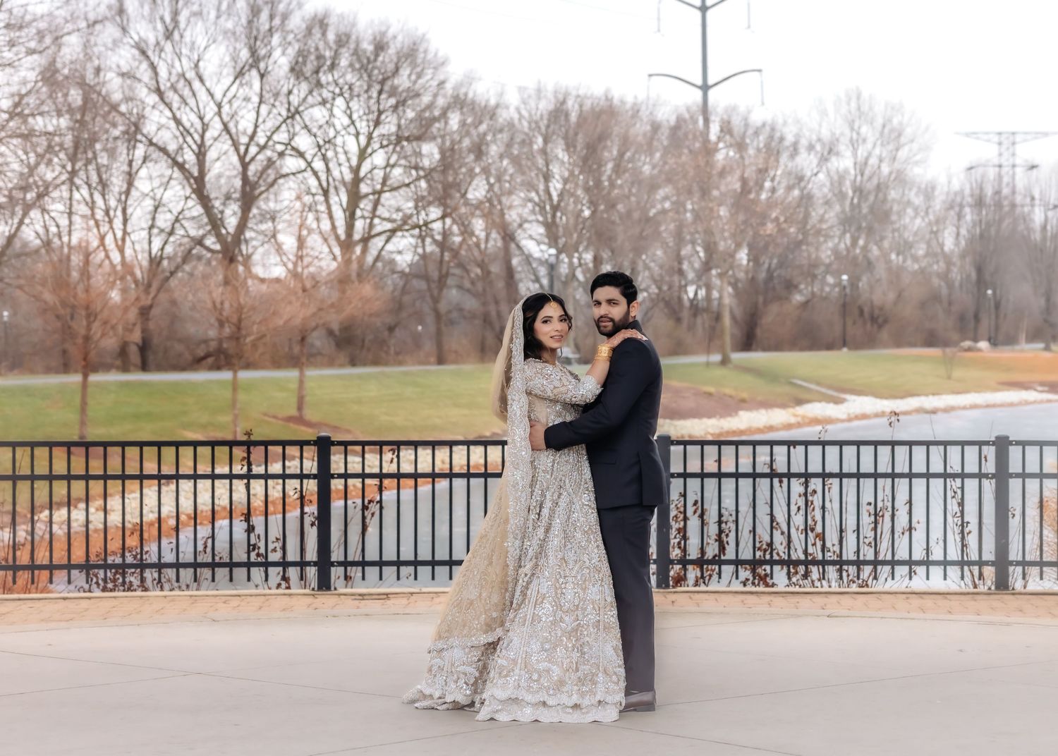 Pakistani wedding couple posing by railing in gold embroidered lehenga and black suit.