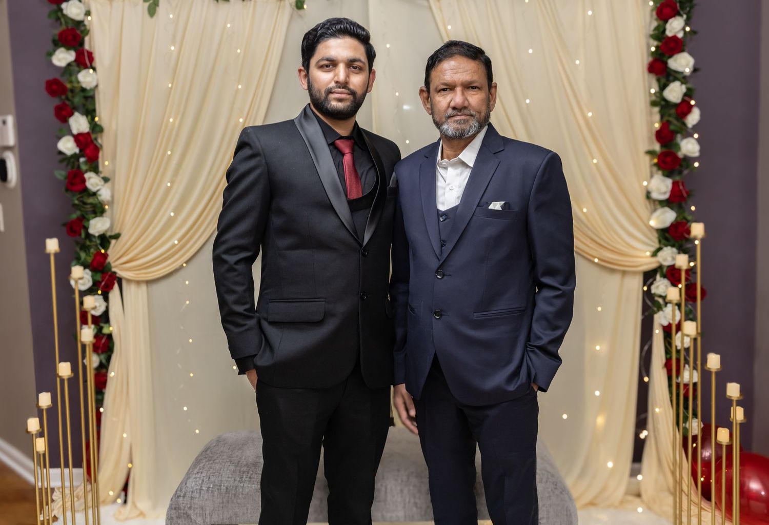 Two men in formal suits pose together in front of a floral wedding backdrop with candle decorations.