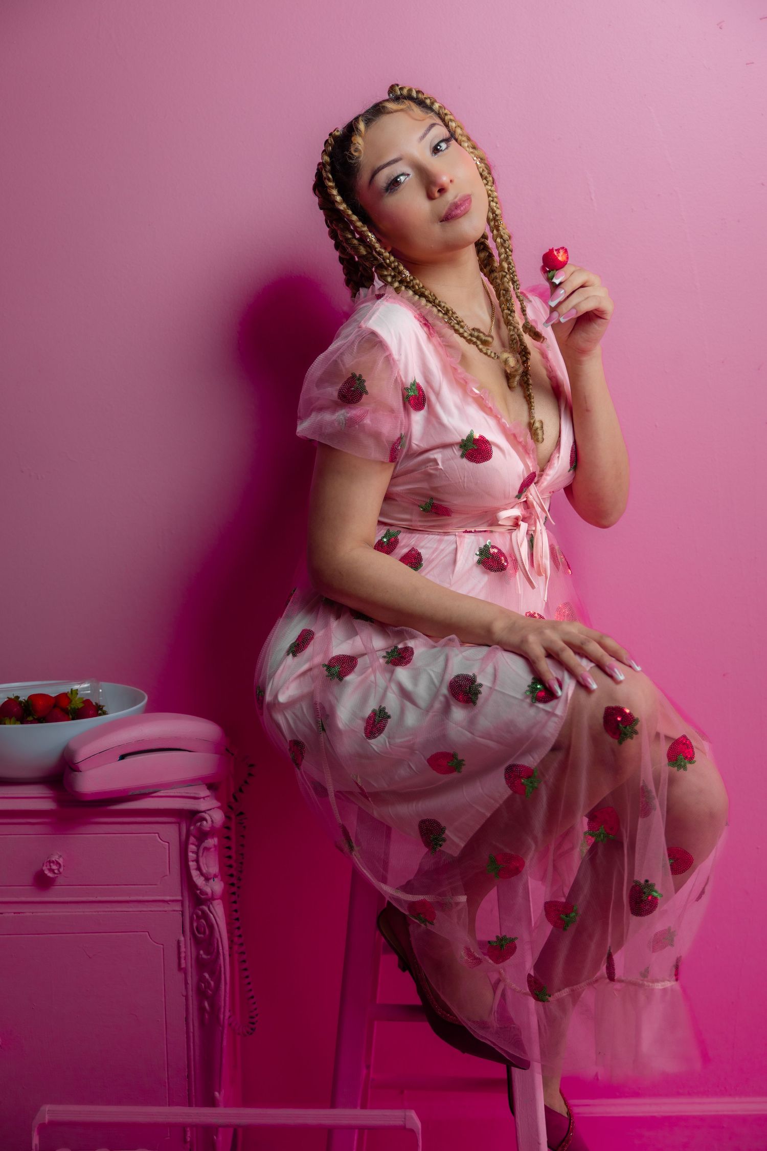 Woman in pink strawberry tulle dress sits on stool holding strawberry against pink wall.