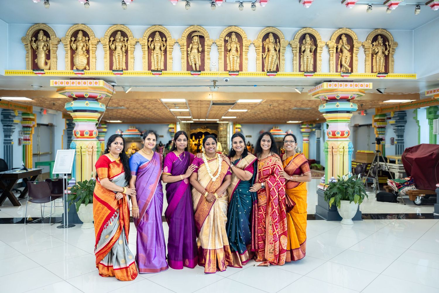 Seven women in elegant colorful silk sarees posing together inside an ornate Hindu temple.