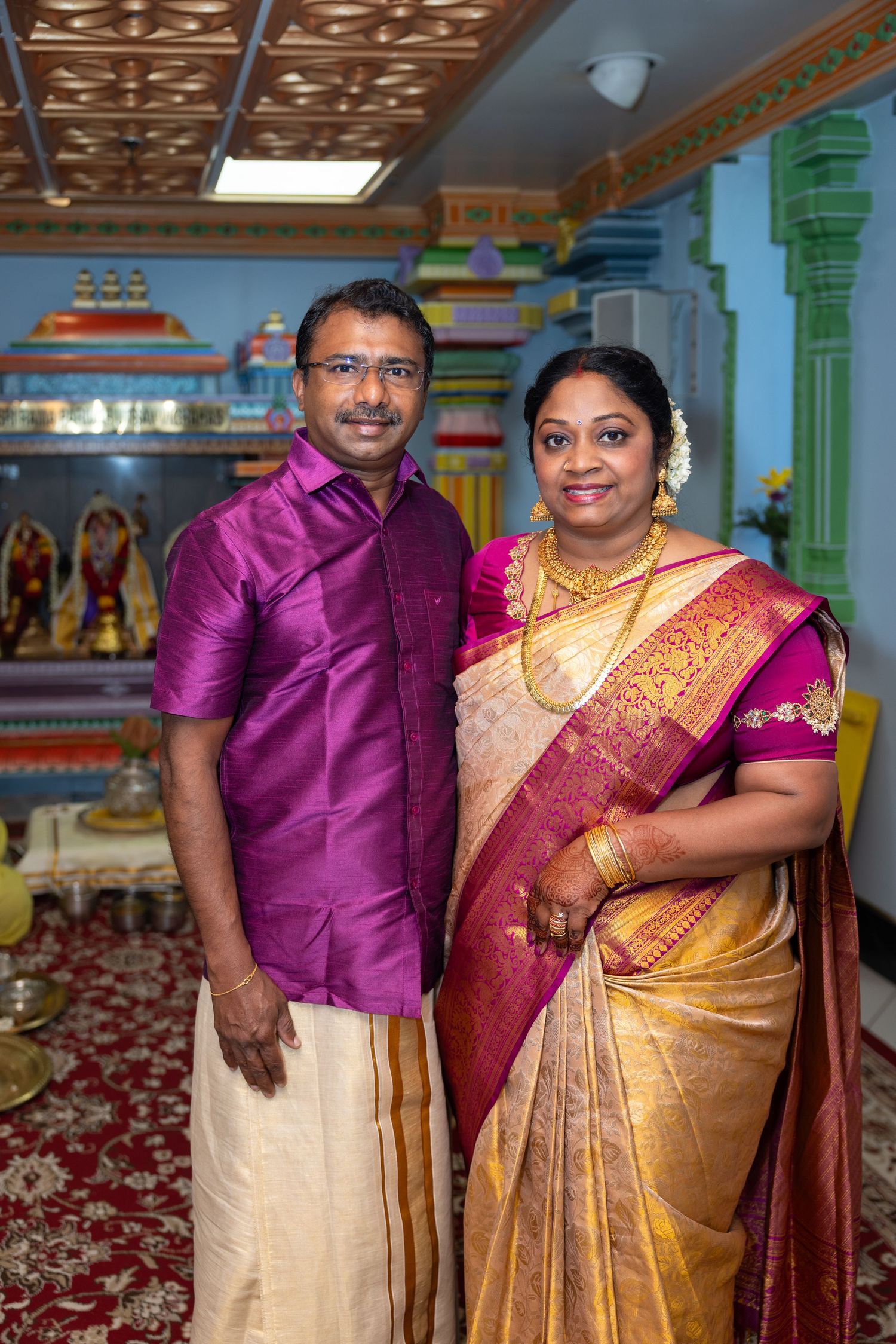 Indian couple in traditional attire, purple kurta and gold saree, pose in decorated temple.