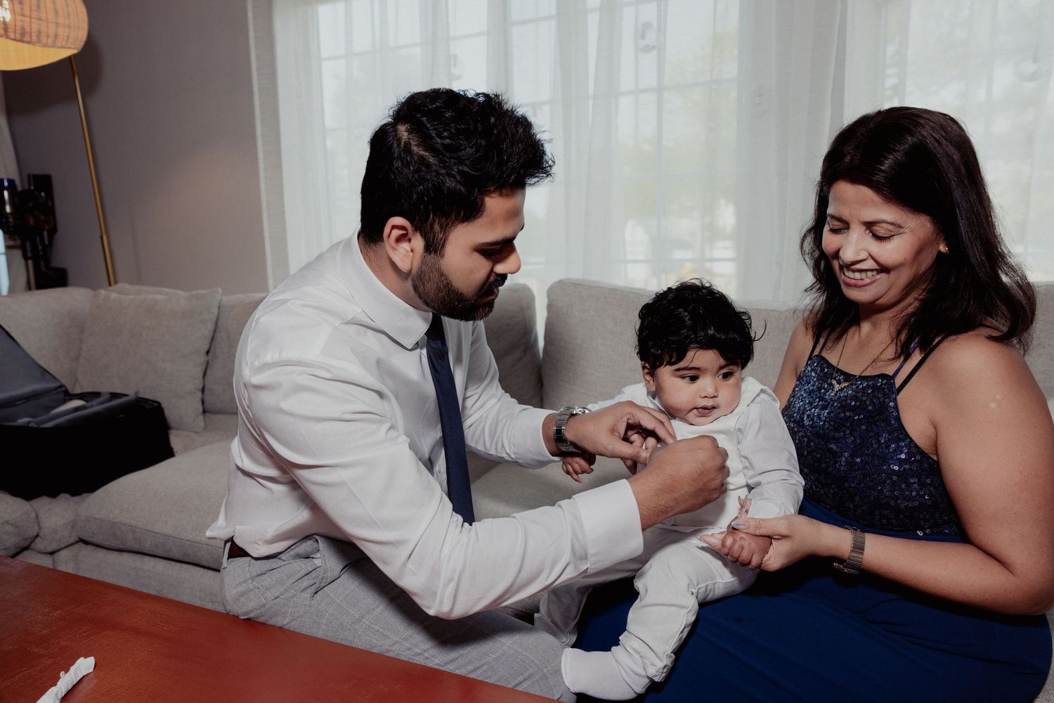 Man in suit helps dress baby held by smiling woman in blue dress on couch.