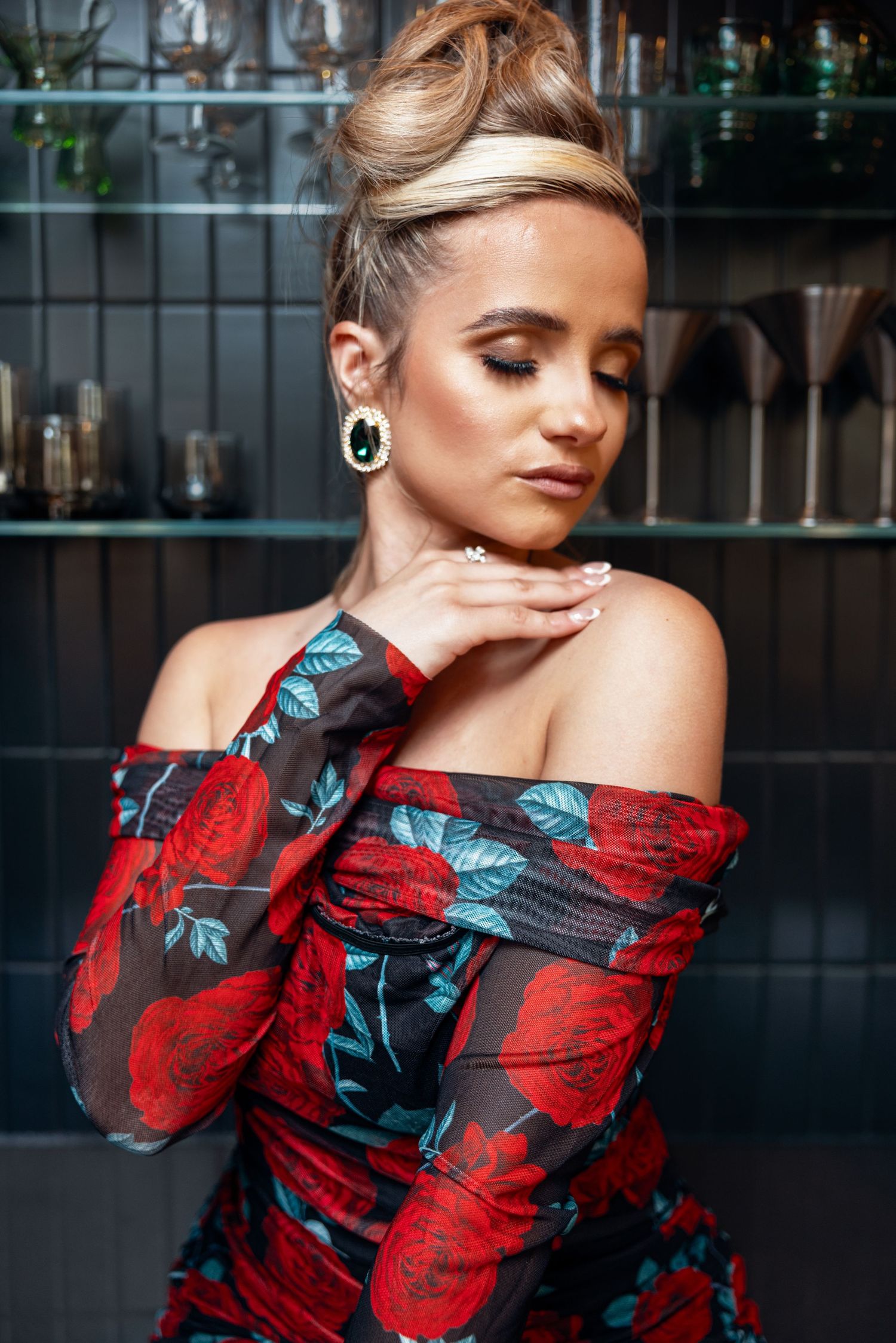 Woman in off-shoulder red rose dress with green earrings posing with eyes closed.
