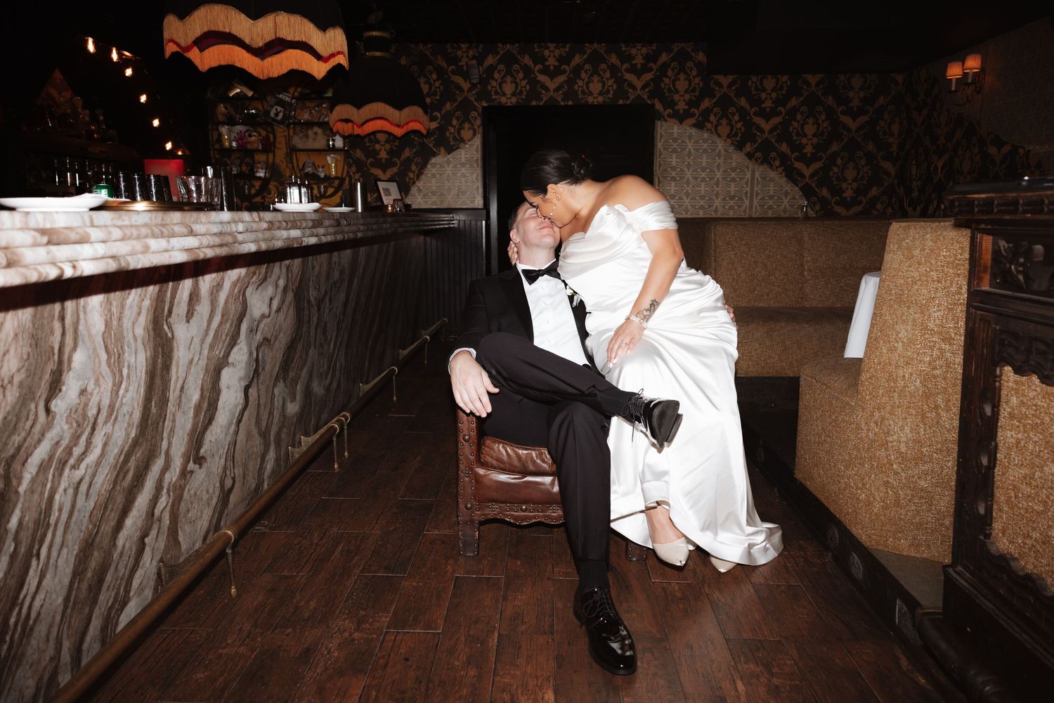Bride sitting on grooms lap sharing intimate kiss in dimly lit rustic bar reception venue.