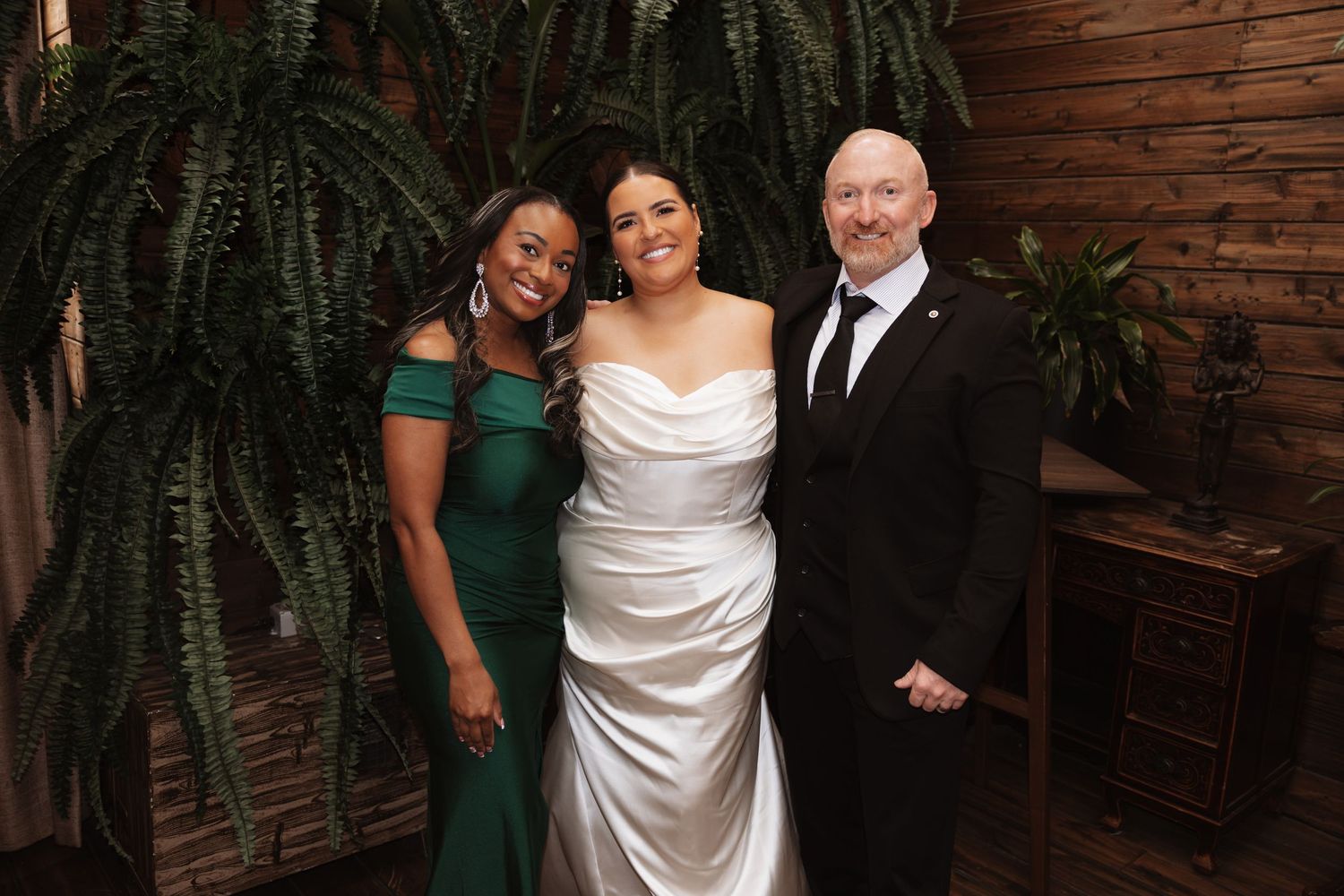 A bride in a white satin gown poses with two guests in front of a lush green tropical plant.