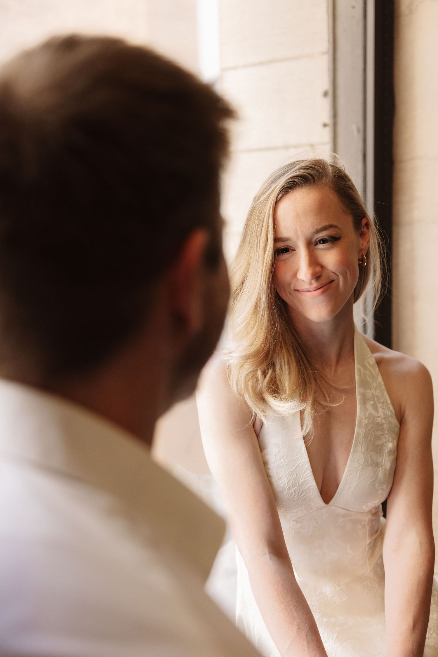 Smiling blonde bride in a deep-V white gown gazes lovingly at her groom in a softly lit interior.