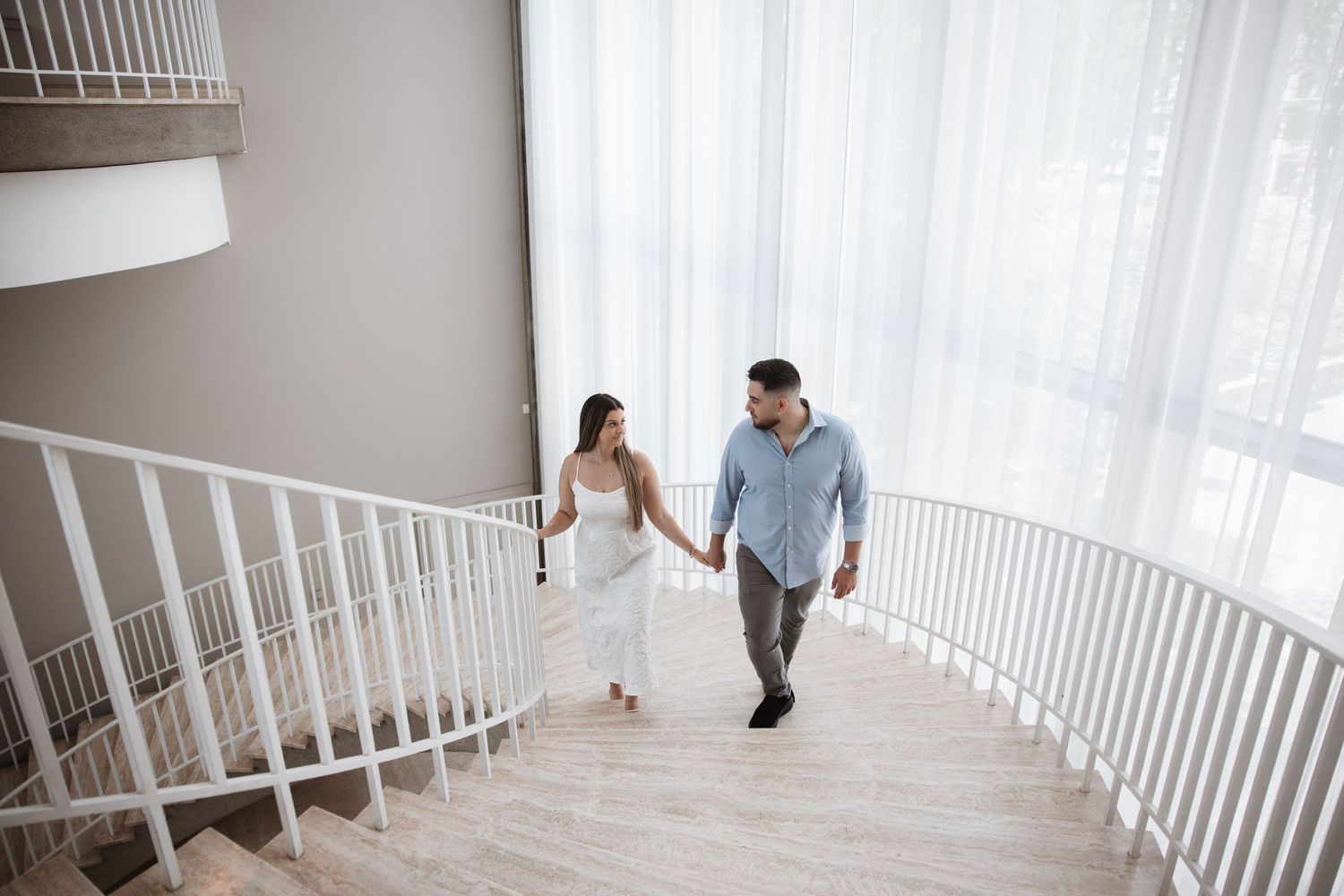 Couple holding hands walking up a modern curved staircase with white railings and sheer curtains.