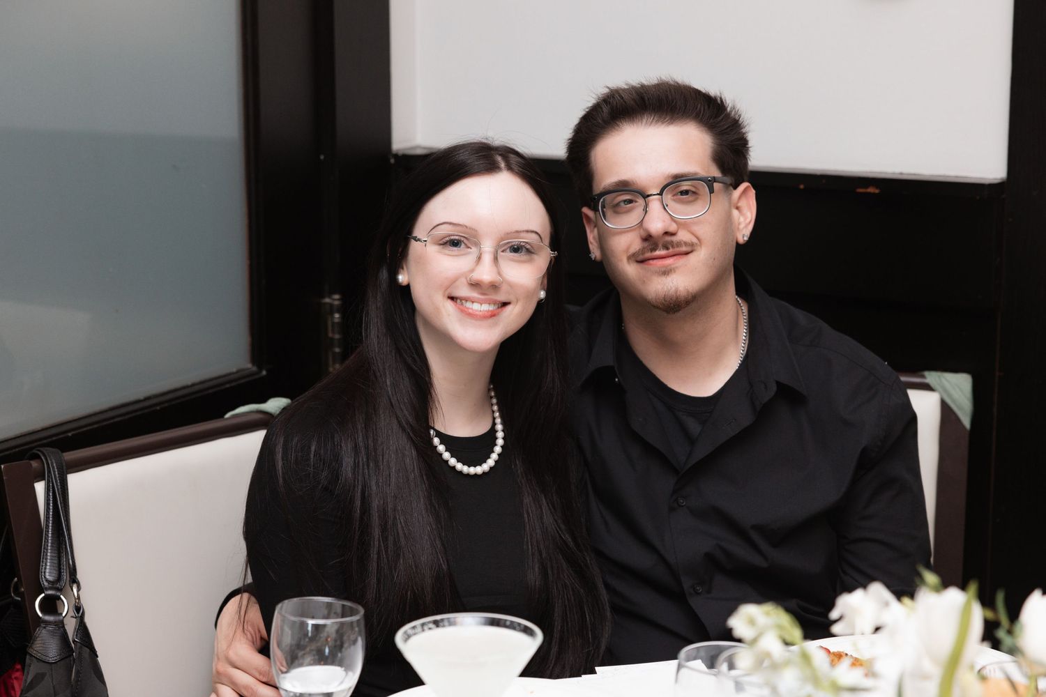 Young couple smiles together at wedding reception dinner table, woman wearing pearls and glasses.