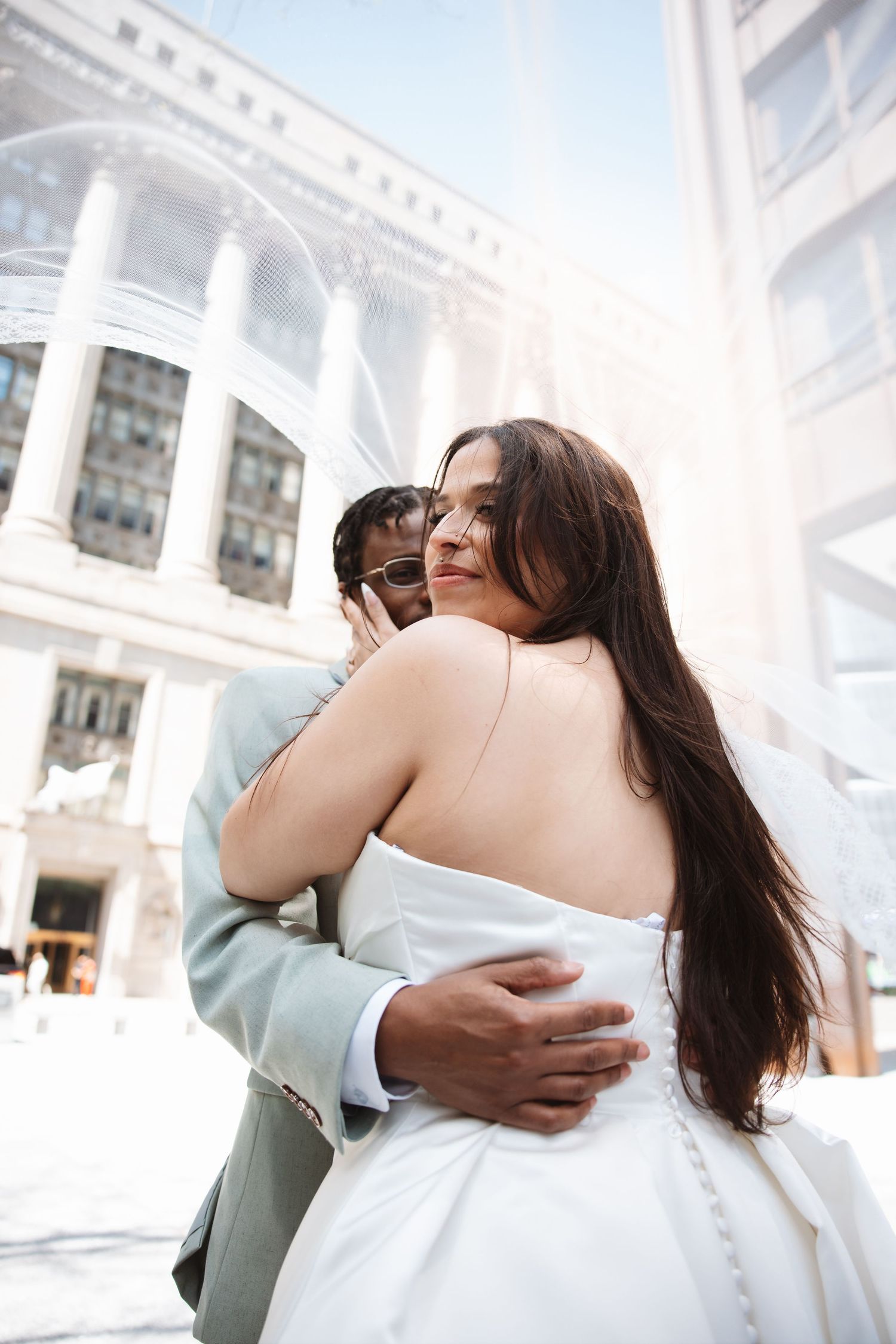 Bride and groom embracing outdoors in front of a grand building, bride in strapless white gown, groom in light gray suit.