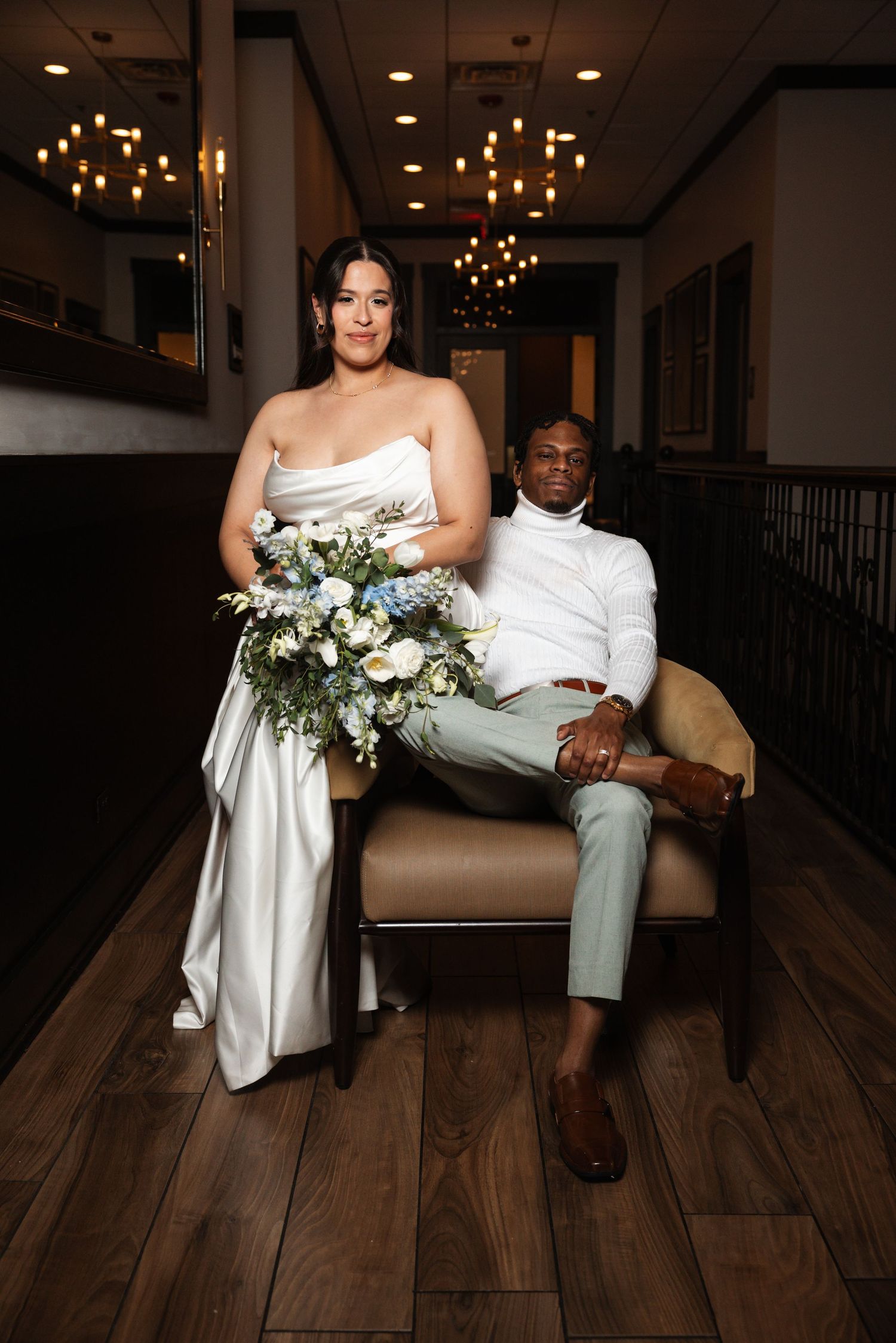 Bride in white strapless gown holds floral bouquet beside groom seated in a chair.