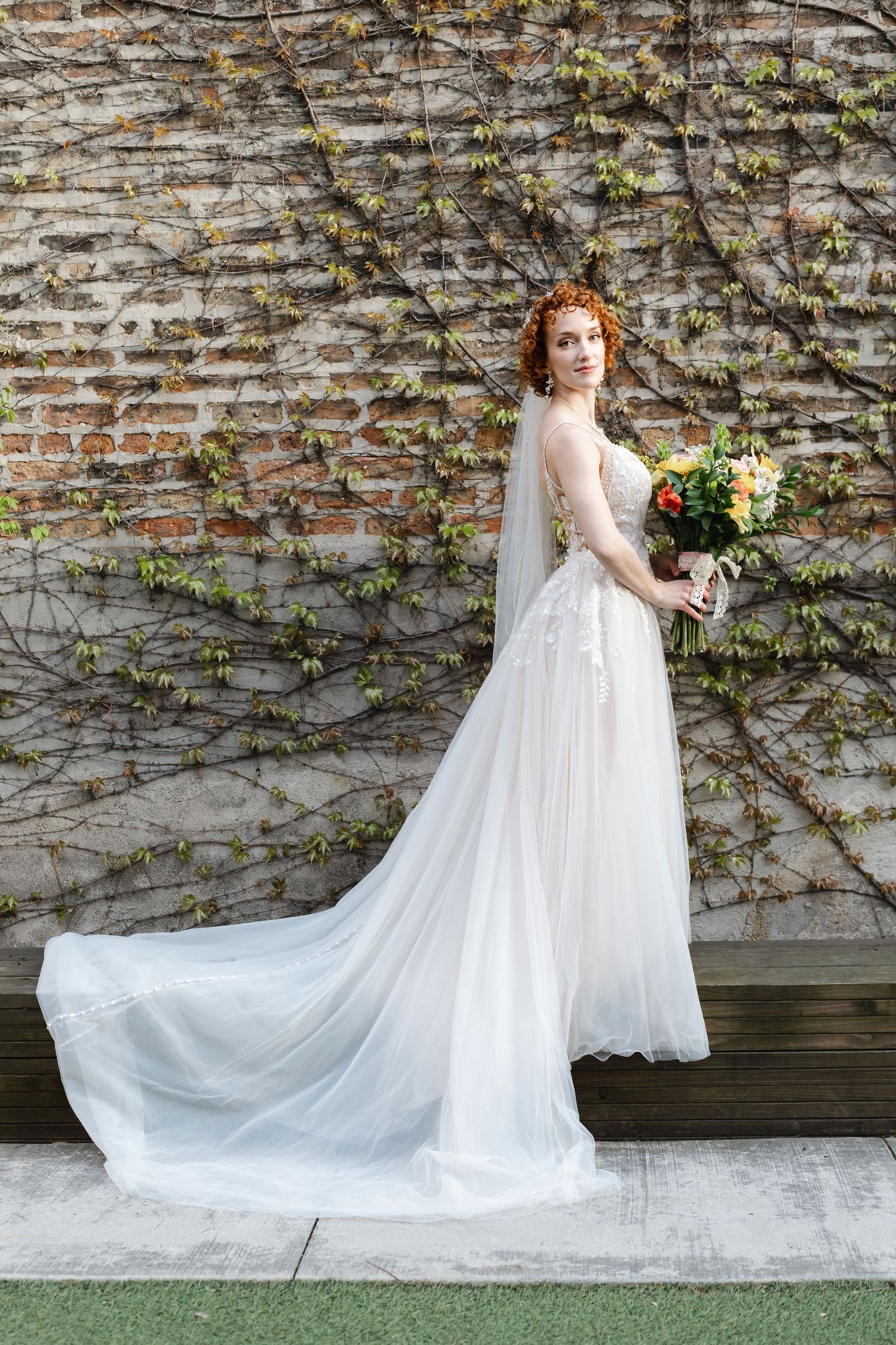 Elegant red-haired bride in a flowing white lace gown with veil holds a bouquet against an ivy-covered brick wall.