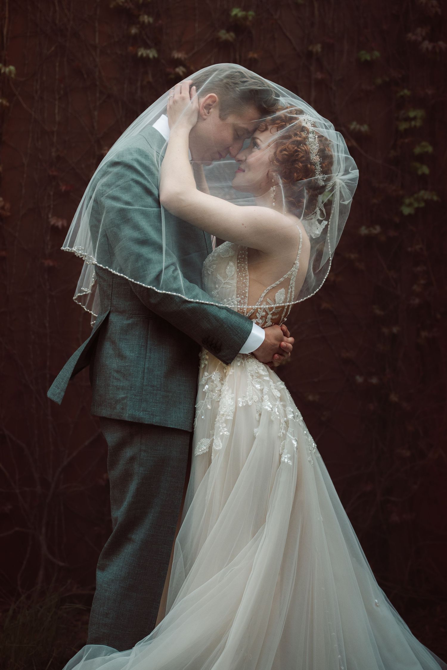 Romantic bride and groom share an intimate moment under a flowing veil against a dark vine-covered wall.