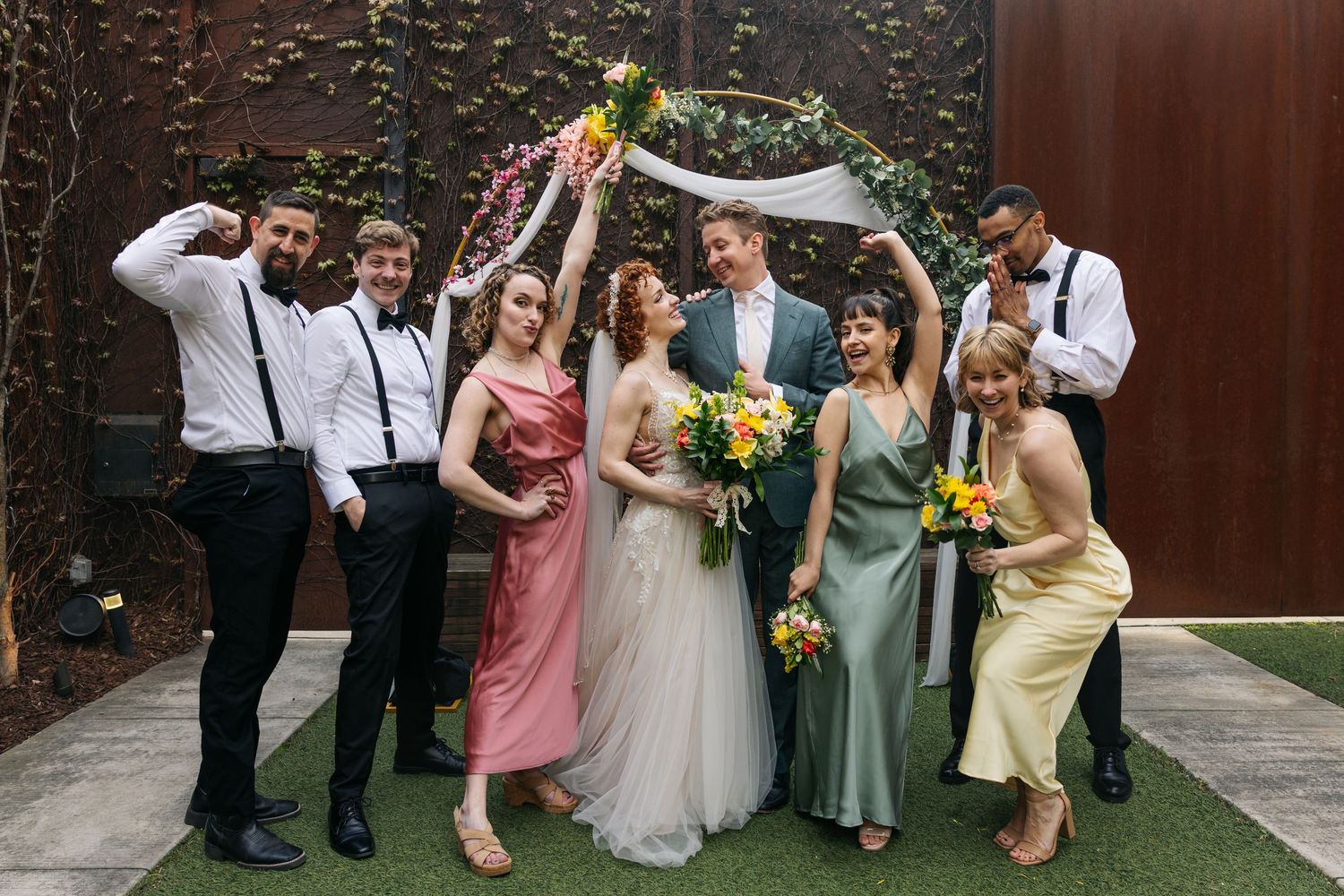 Wedding party poses joyfully under a floral hoop arch, bridesmaids in colorful dresses with groomsmen in suspenders.