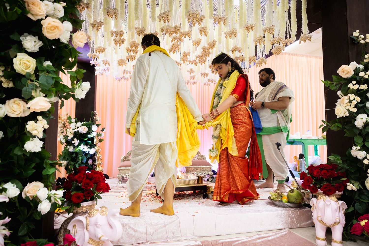 Indian bride in red saree and groom in white kurta walk together at decorated Hindu wedding mandap.