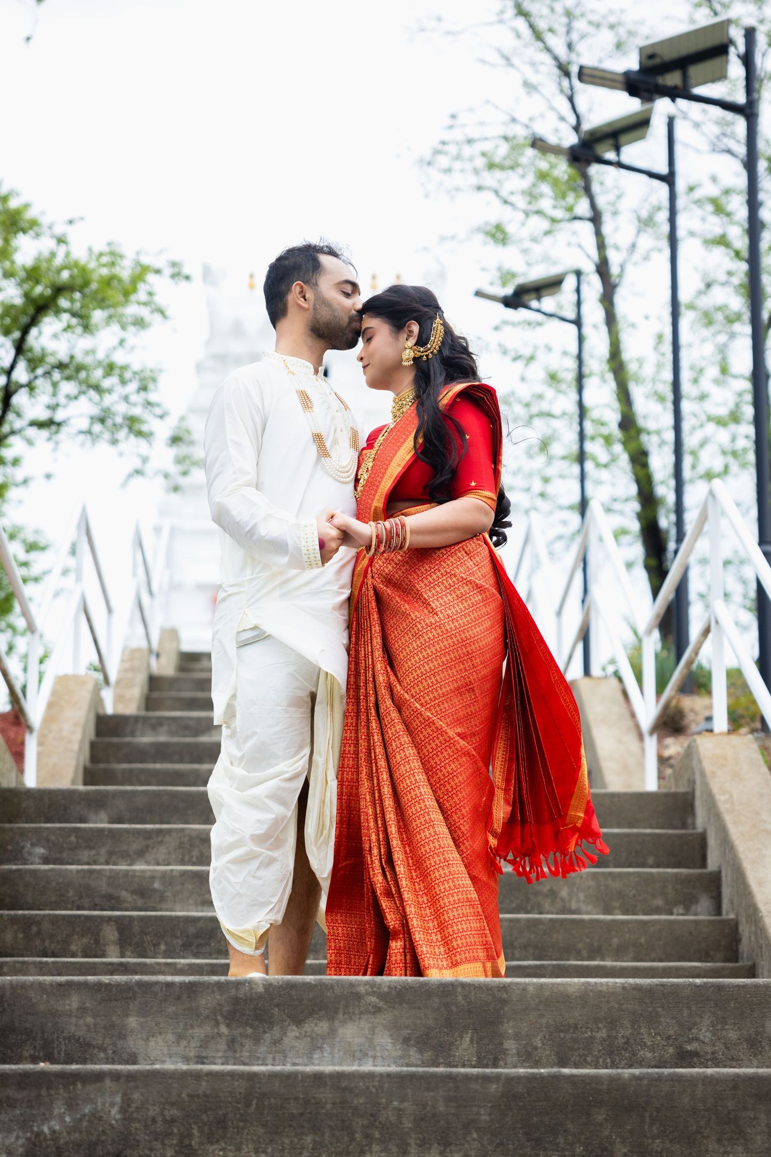 Indian groom kissing bride on temple steps, bride wearing red silk saree and gold jewelry.