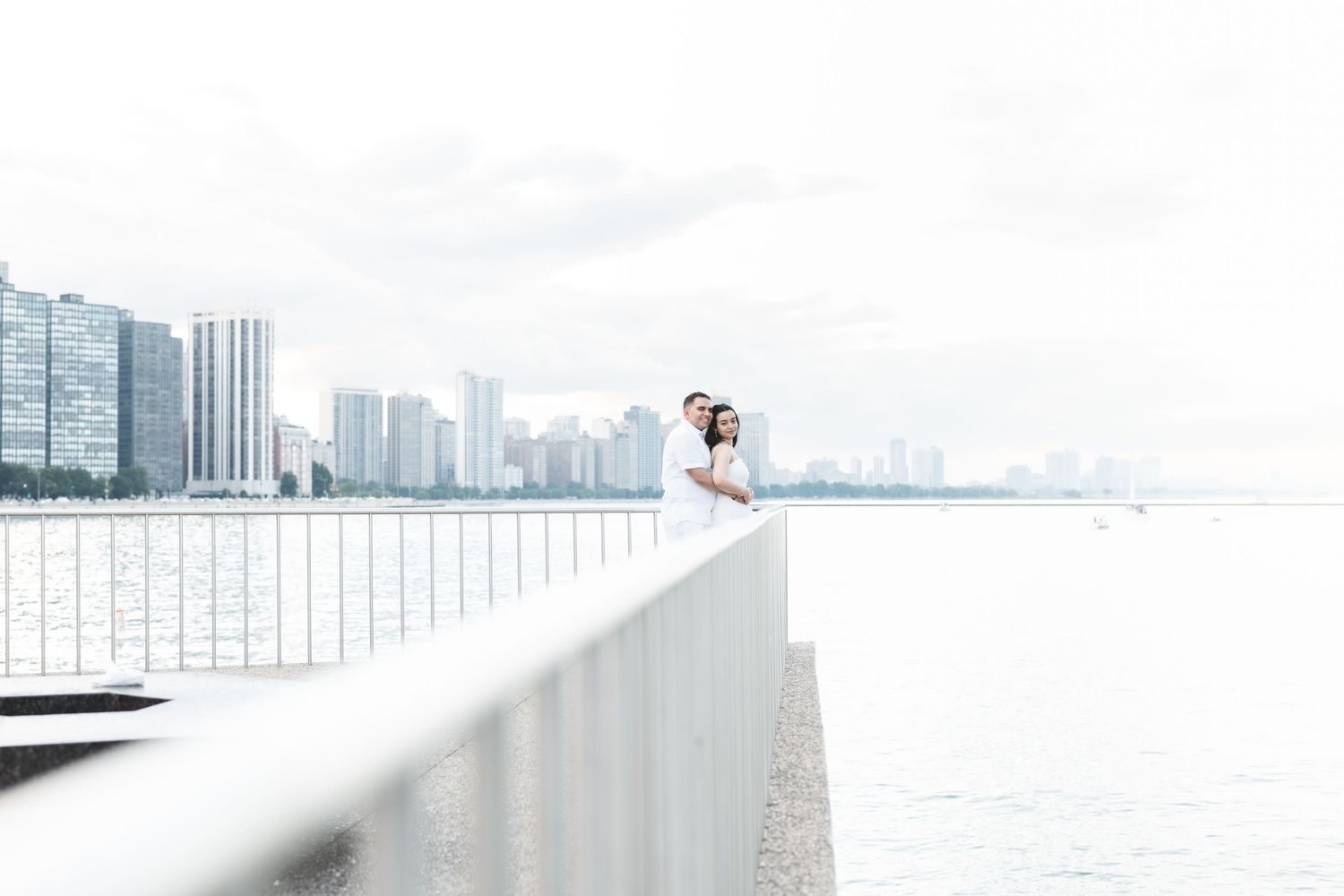 A couple in white standing on a pier with a bright city skyline and lake view.