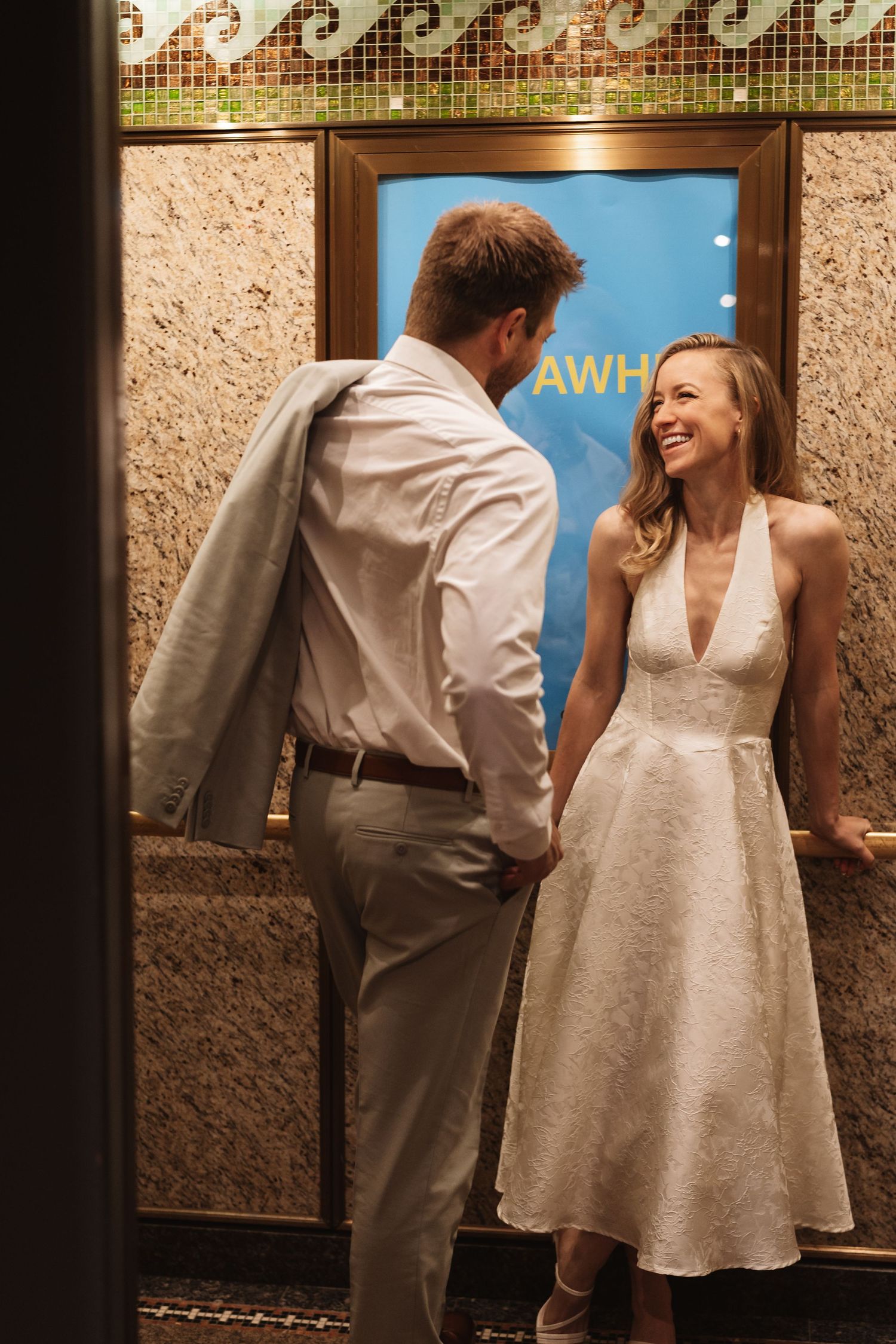 Bride in white halter dress laughs with groom in white shirt outside a venue.