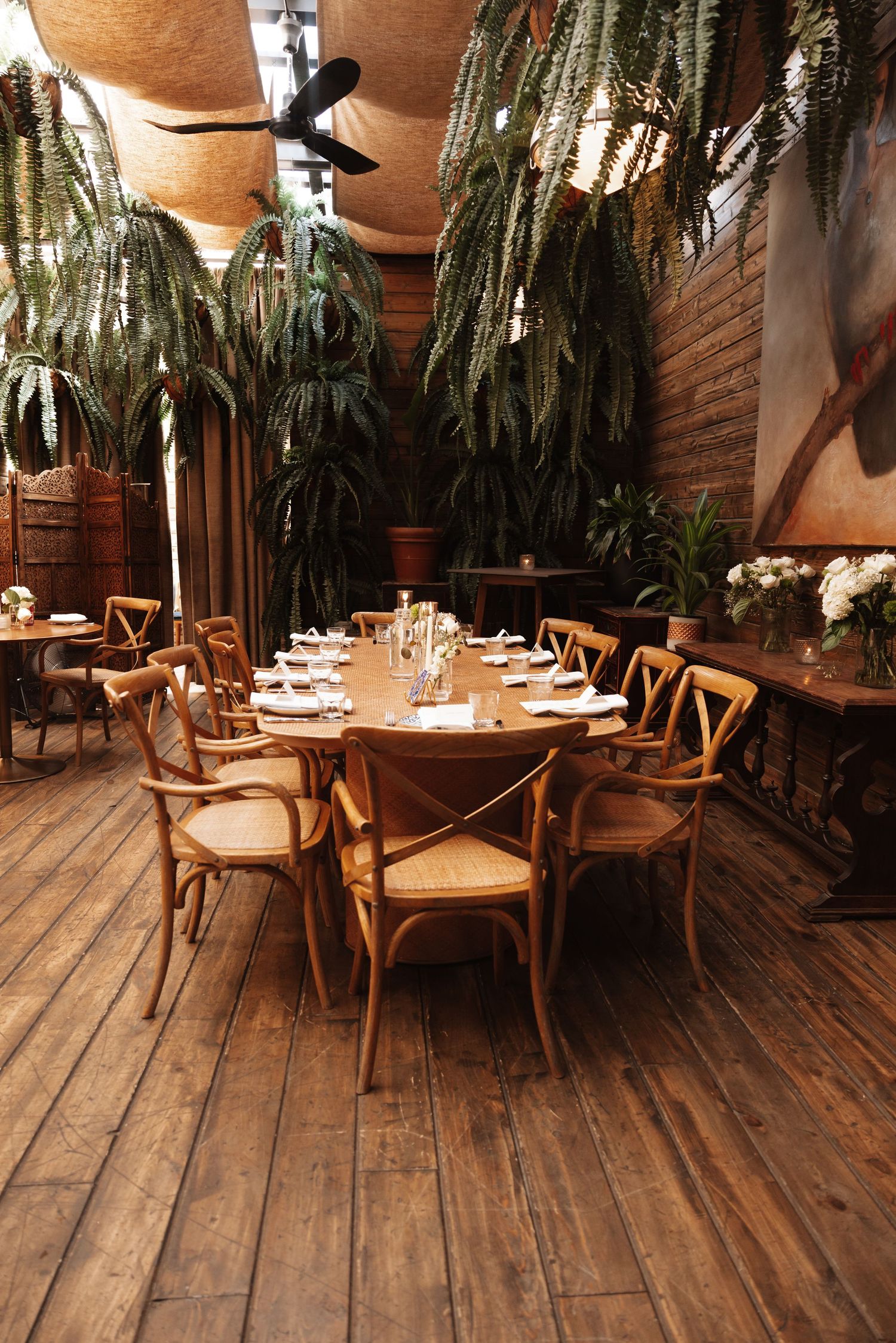 Elegant rustic wedding reception venue with round wooden tables, cross-back chairs, and lush tropical plants.