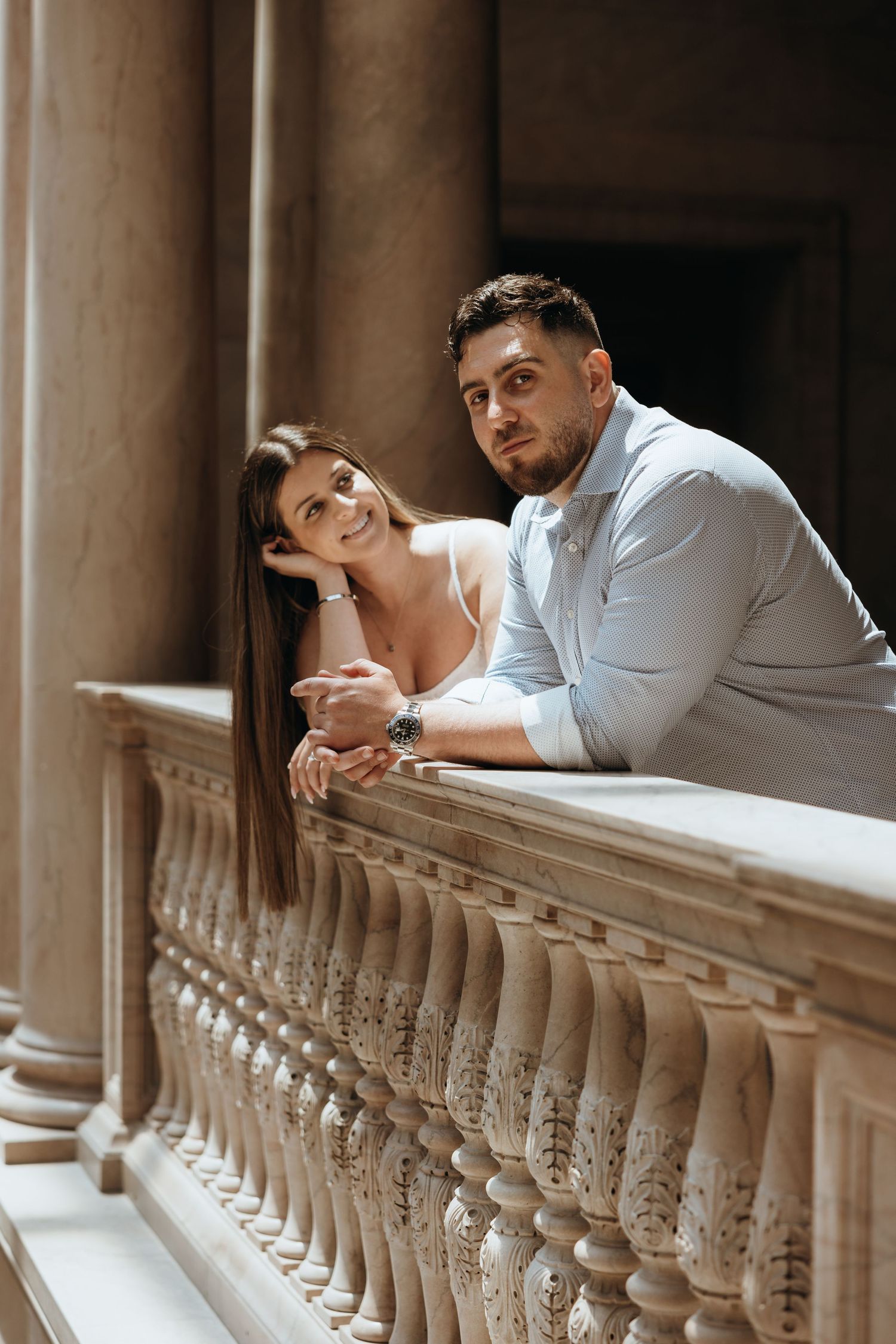 Smiling couple leaning on decorative stone balcony railing between tall columns in a classical building.