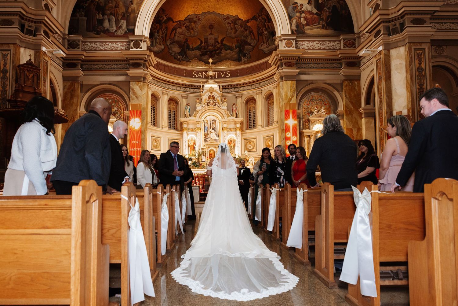 Bride walks down the aisle in a stunning white gown at an ornate Catholic church wedding ceremony.