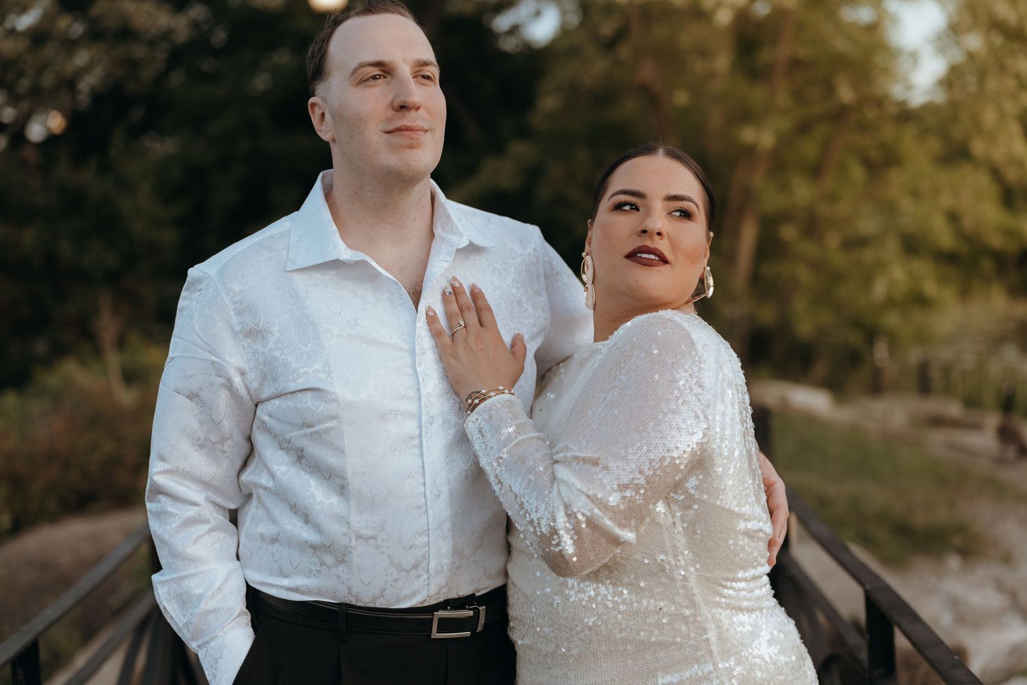 Bride in a sparkling gown poses with groom on a bridge during outdoor wedding portraits.