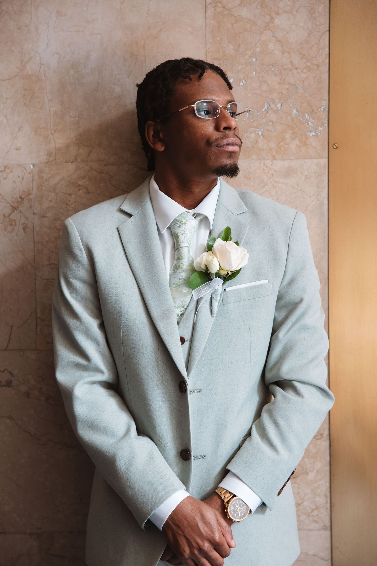 Elegant groom in light gray suit with boutonniere and tie, wearing glasses, posing against marble wall.