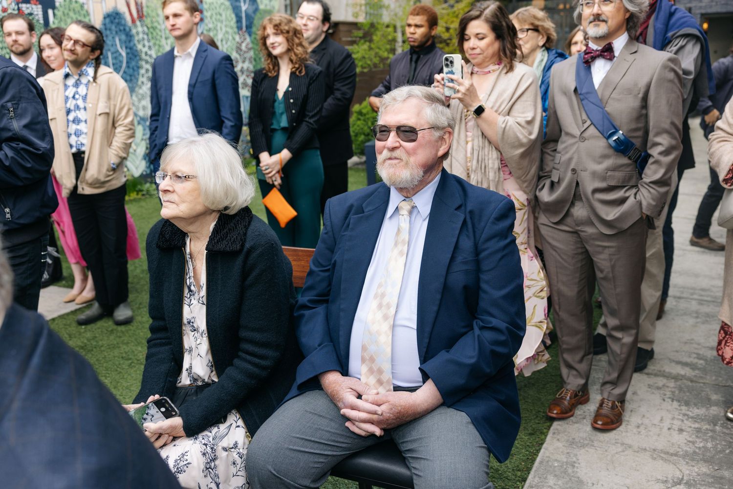 Elegantly dressed guests seated and standing outdoors at a formal gathering or reception event.