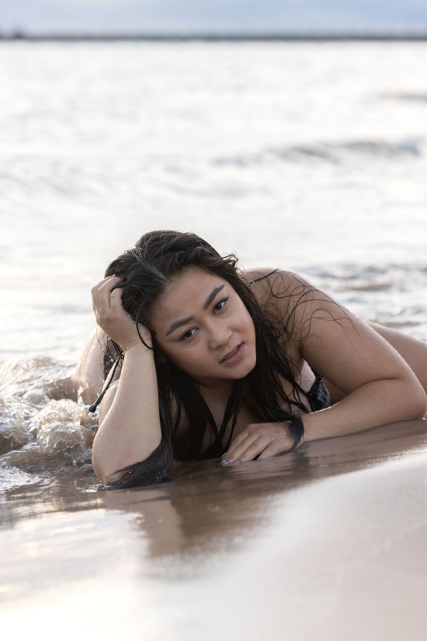 Young woman with wet hair lying in shallow ocean waves on a sandy beach.