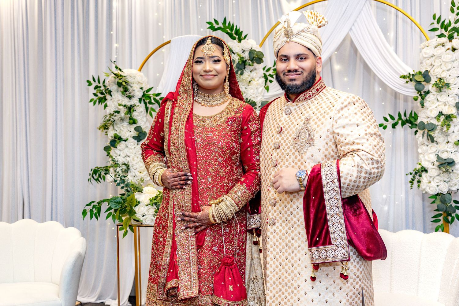 South Asian bride and groom smiling in traditional red and gold wedding attire before white floral backdrop.