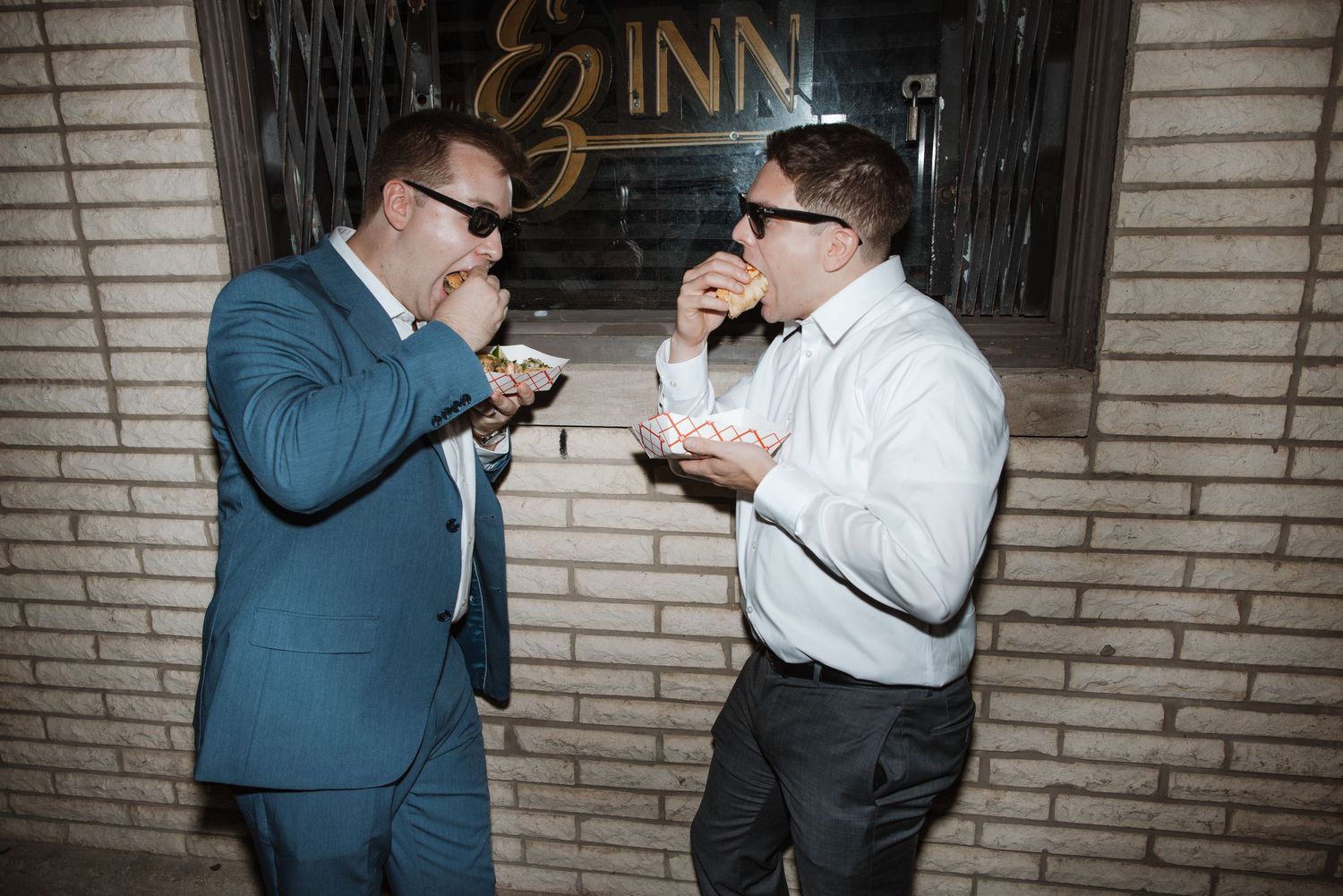 Two men in suits eating and laughing outside an inn at night.