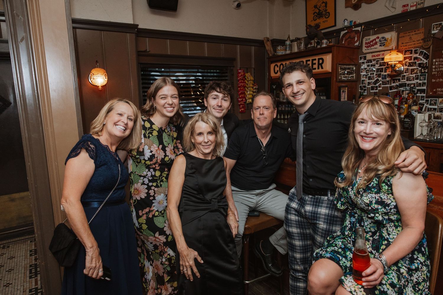 Group of seven wedding guests smiling together inside a warmly lit restaurant bar.